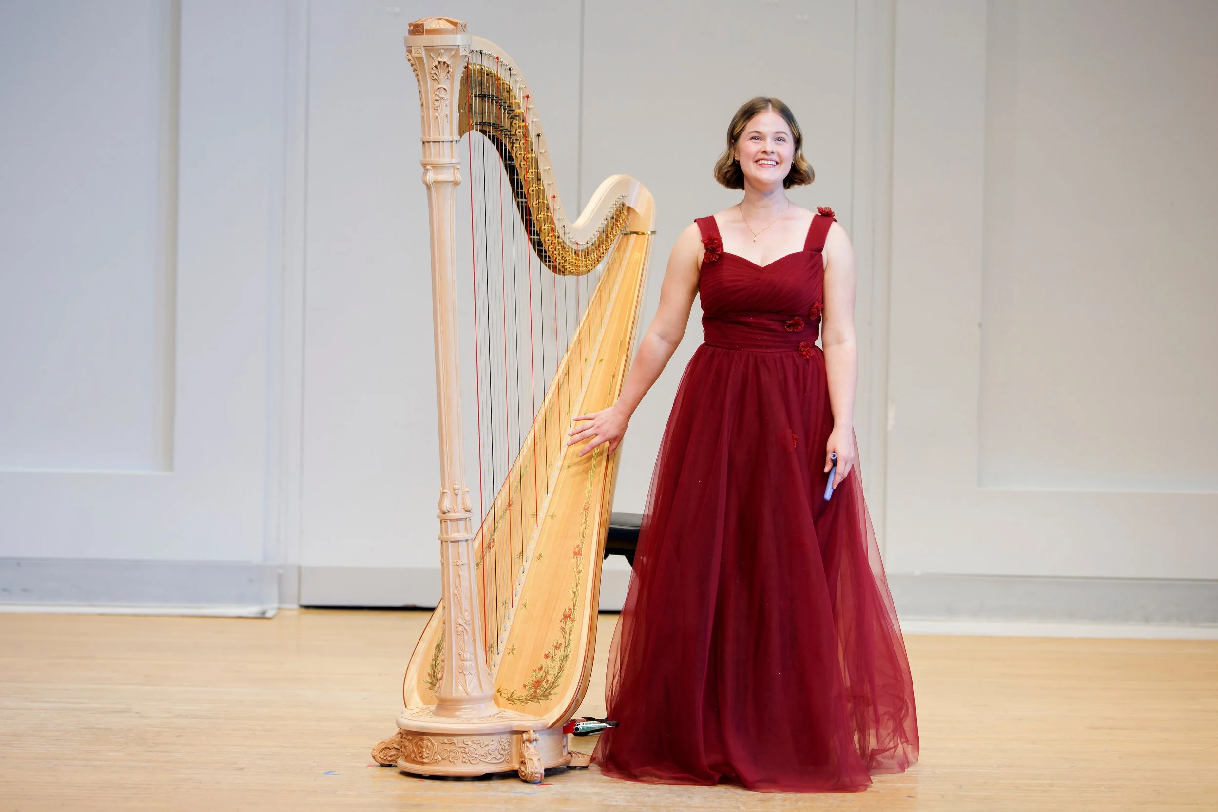 A woman in a red gown standing next to a large wooden harp in a performance setting.