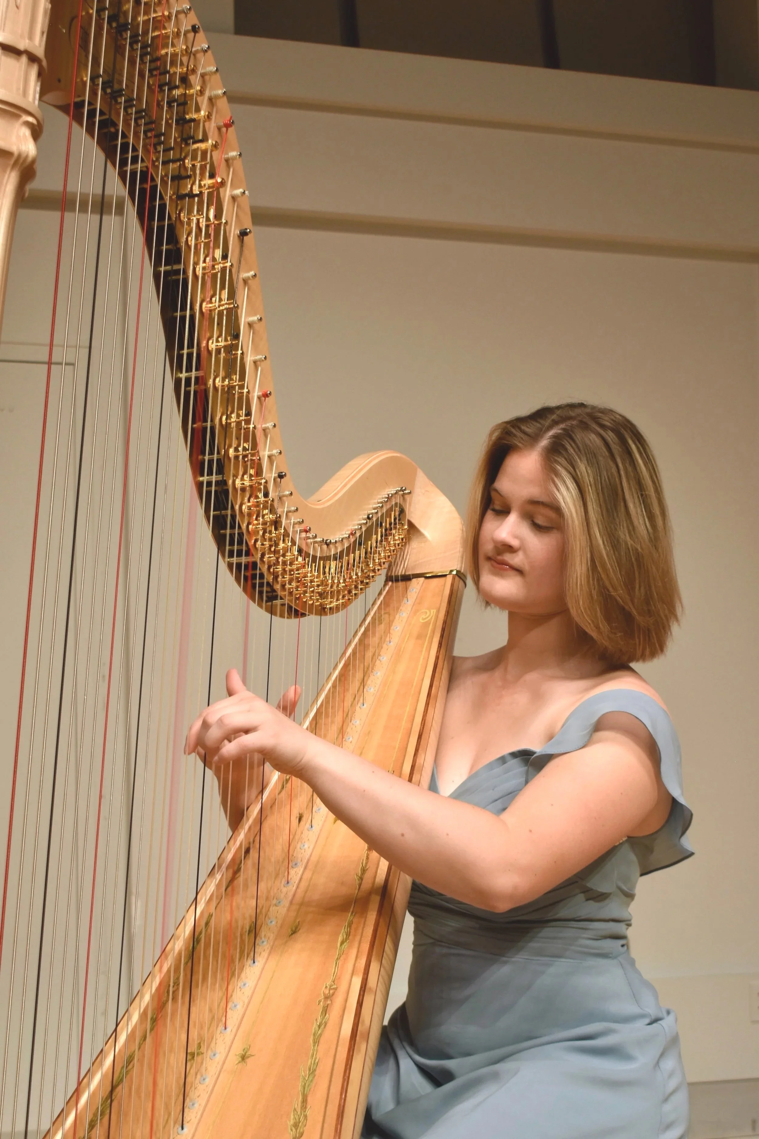 A woman in a gray dress playing a large wooden harp in a room with cream-colored walls.