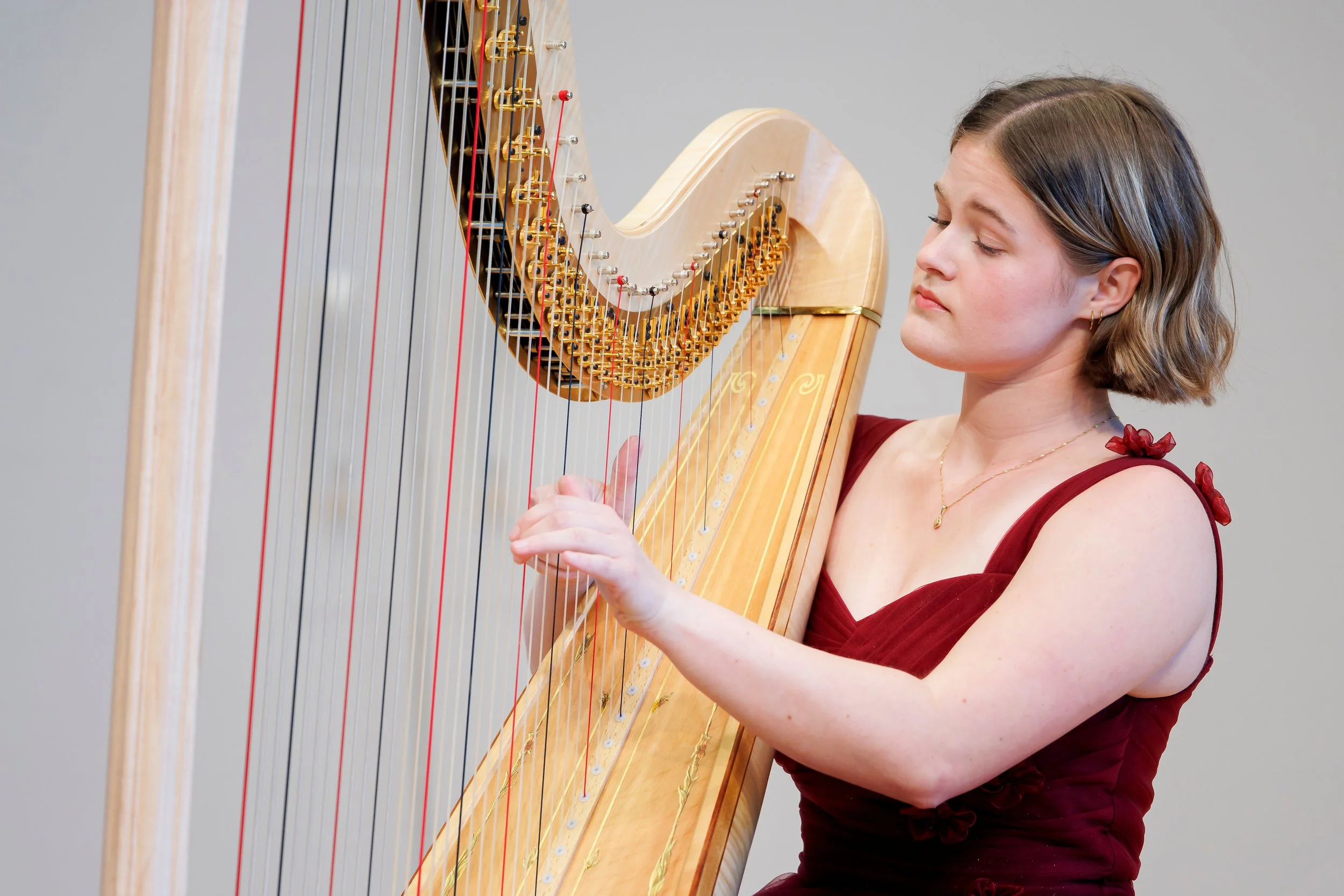 A woman playing a harp with her eyes closed, wearing a red dress, in front of a gray background.