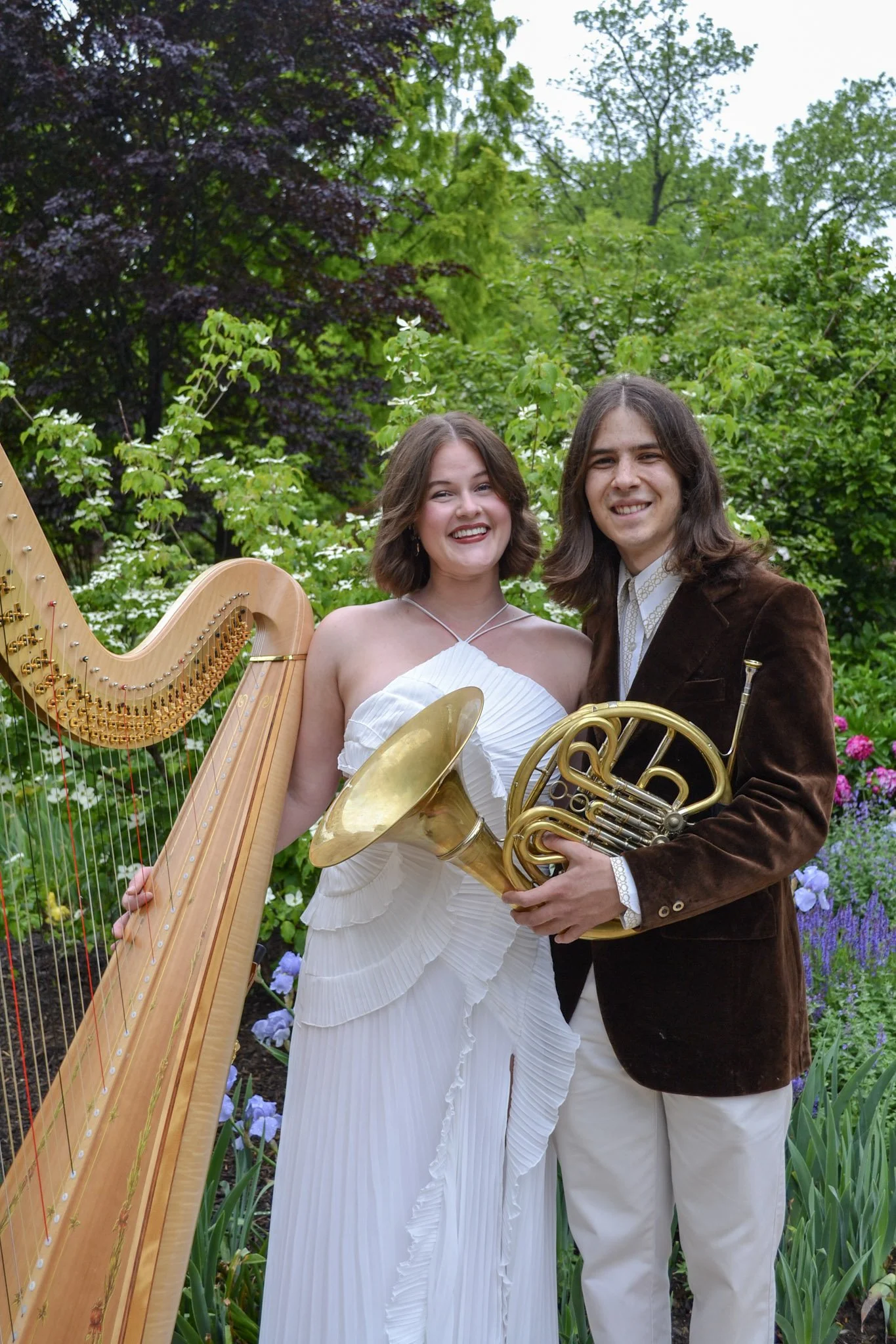 A man and woman smiling outdoors, holding musical instruments, in a garden with green plants and flowers.