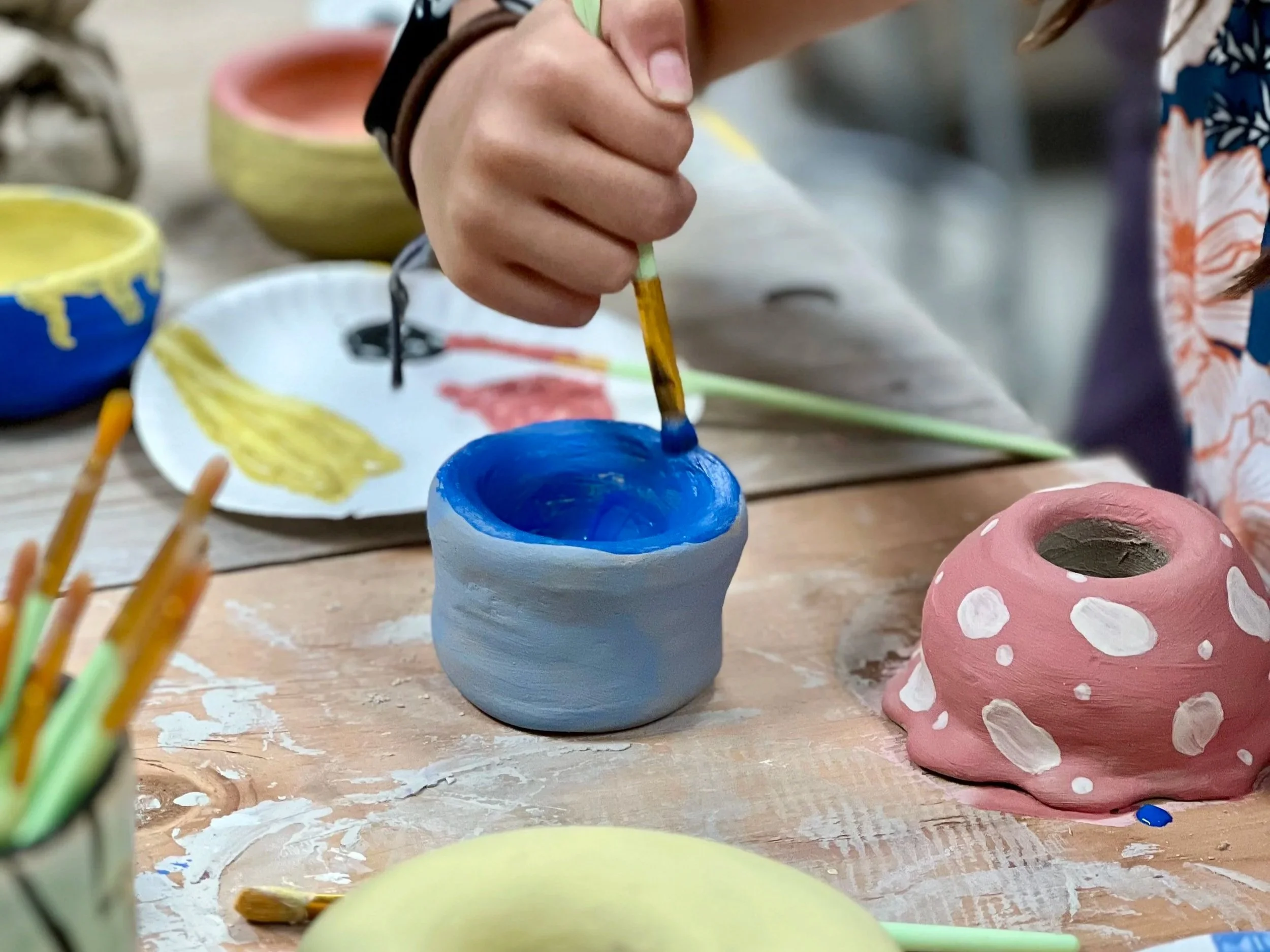 A child's hand paints a ceramic vessel with blue underglaze