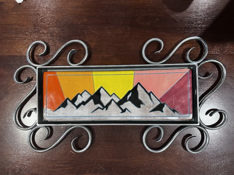 A hand-made ceramic tile with a mountainscape on a colorful background surrounding be an ornate metal frame.