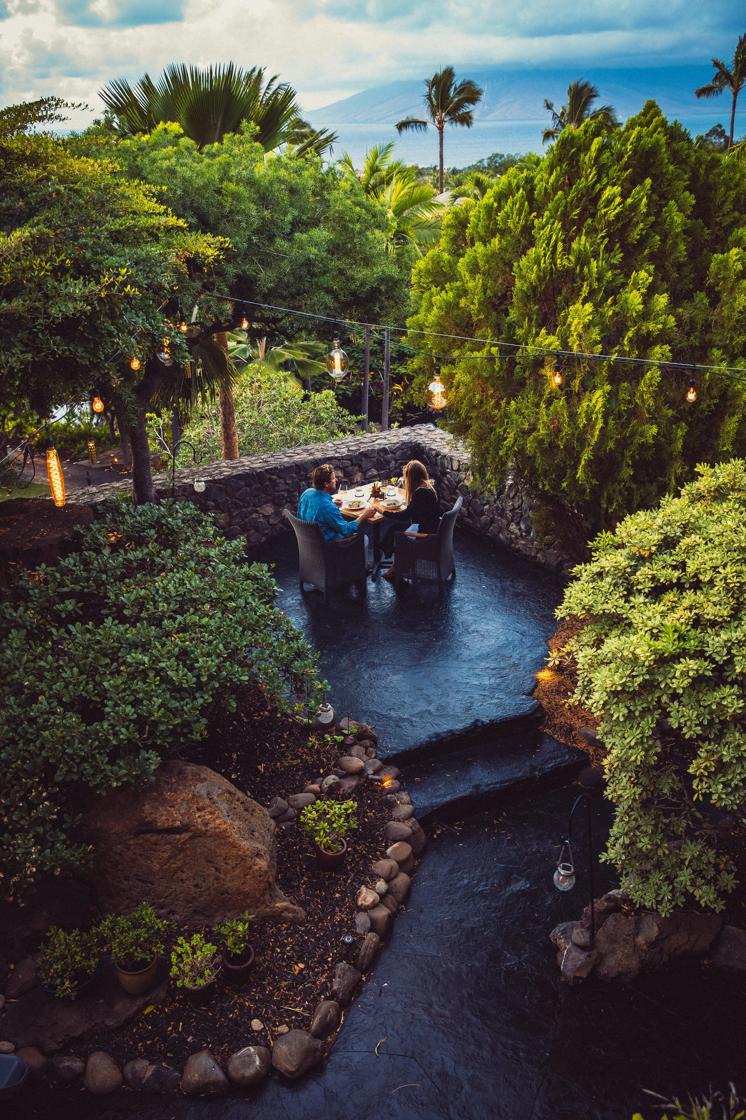 People dining outdoors on a patio surrounded by lush greenery, with string lights hanging above, overlooking a scenic view of a volcano and ocean in the distance.