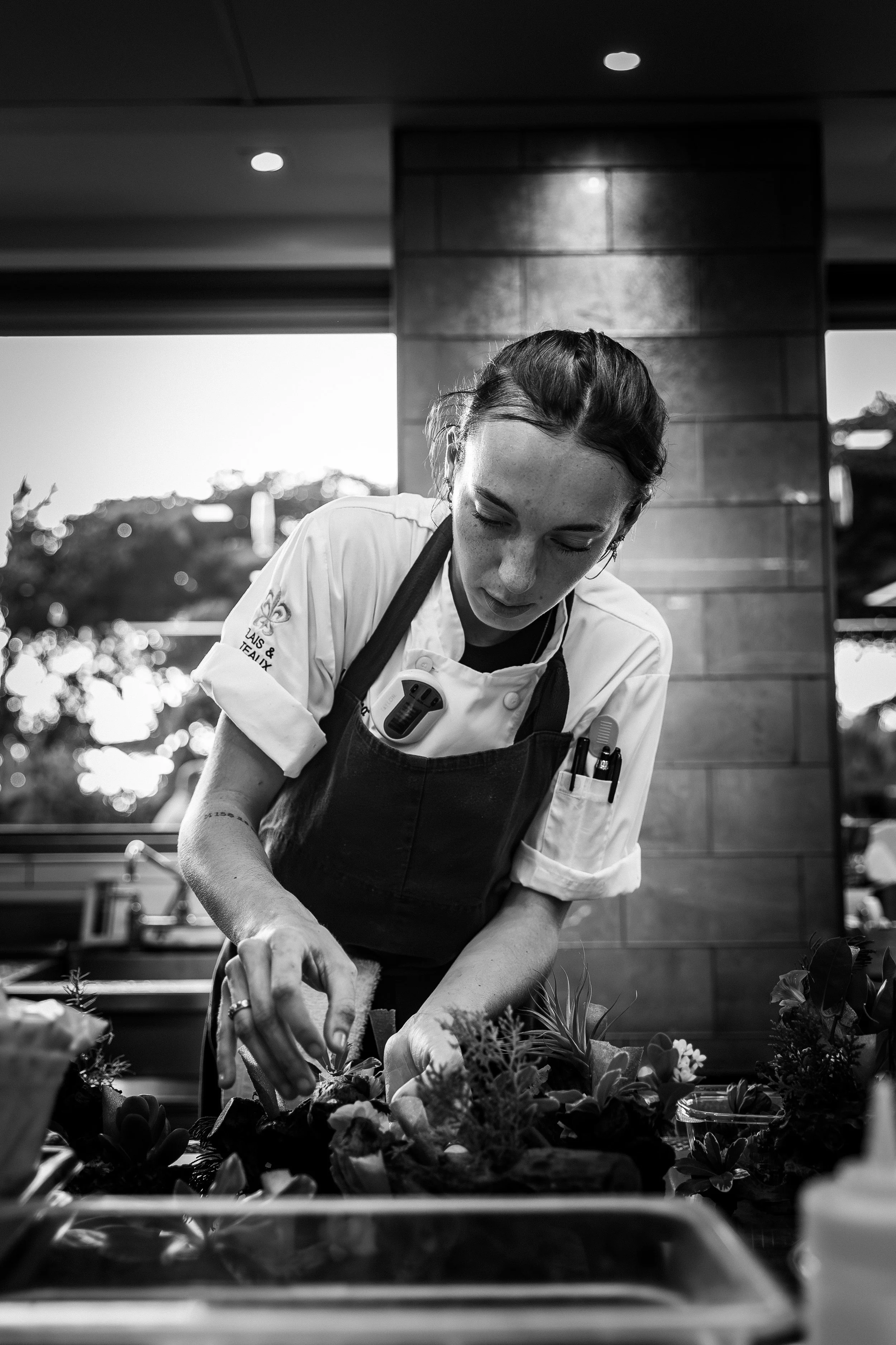 A female chef in a white coat and apron preparing a dish with vegetables in a kitchen.