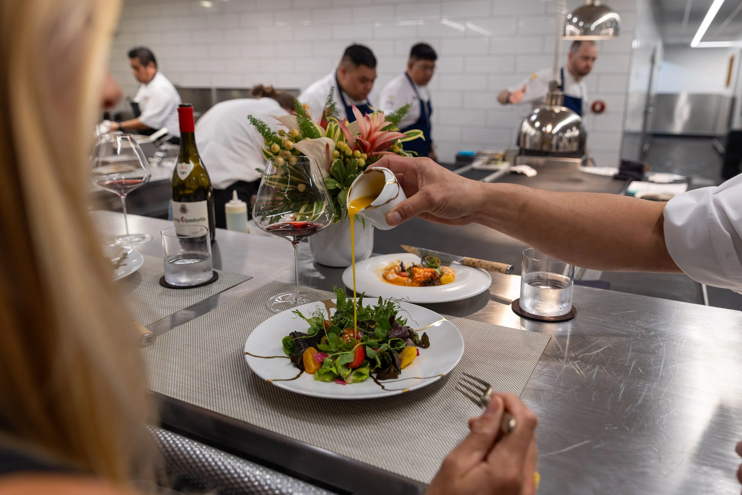 A person pouring dressing over a salad in a restaurant kitchen with chefs working in the background.