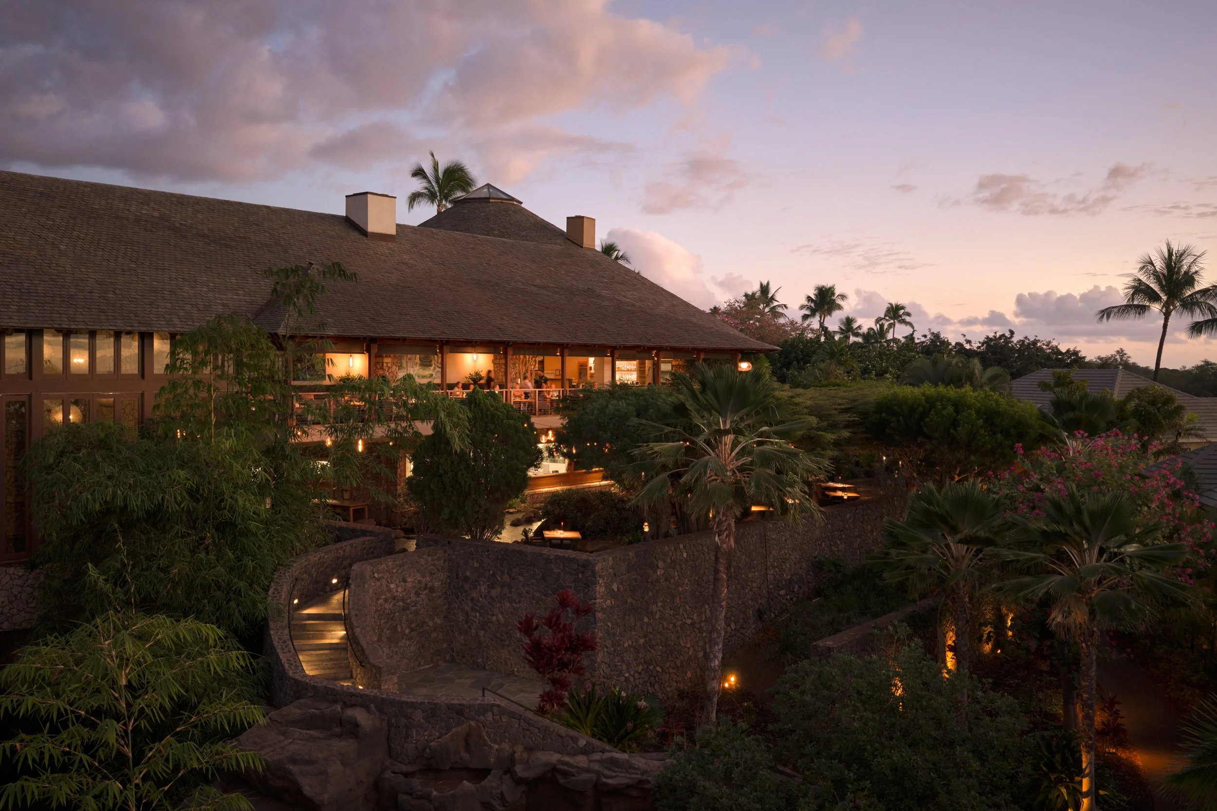 A large house with a sloped roof surrounded by palm trees and lush greenery at sunset, with warm lights glowing from inside.