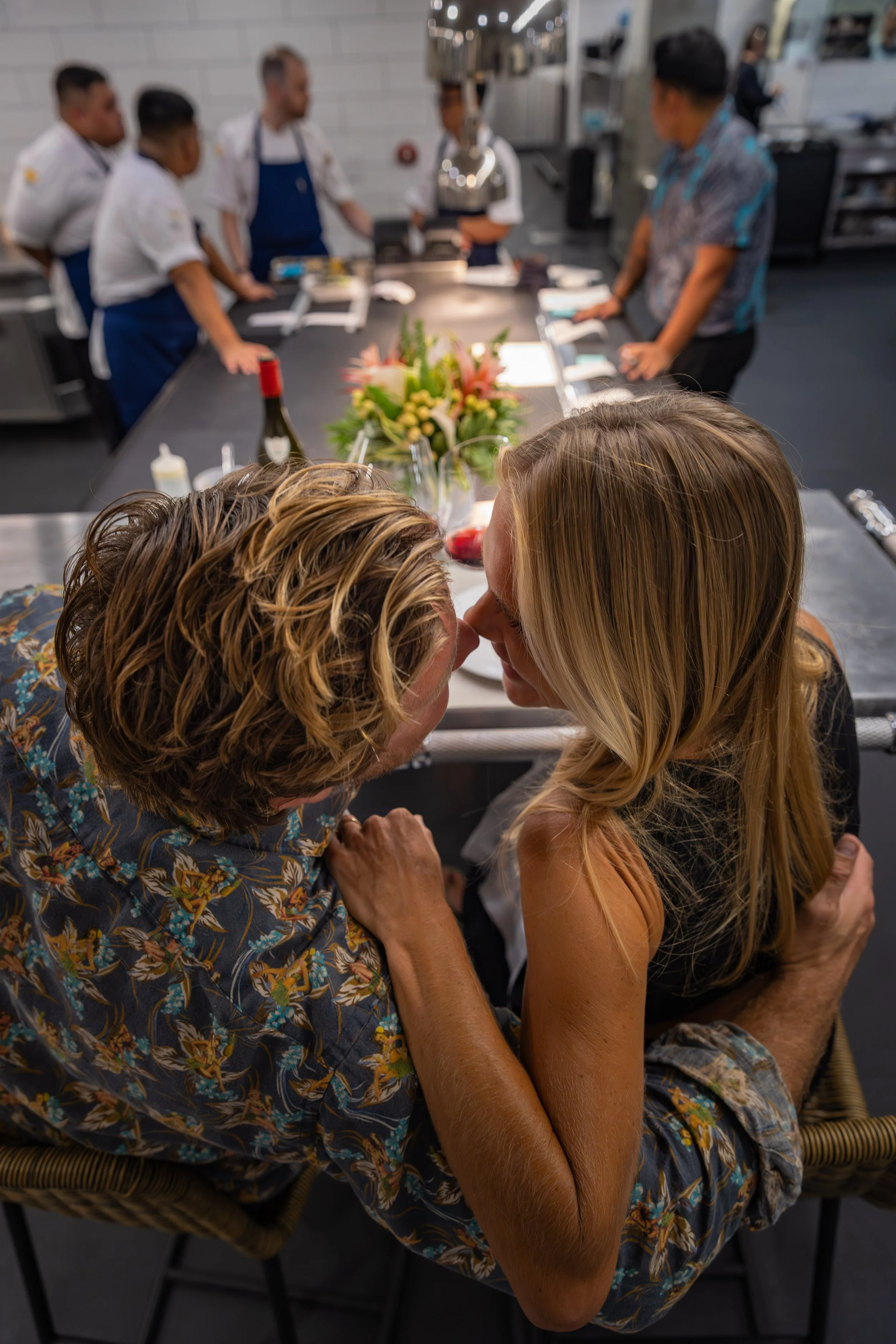 A couple is sharing a kiss at a table with a floral centerpiece, wine, and glasses, in a restaurant or kitchen setting with chefs and kitchen staff in the background.