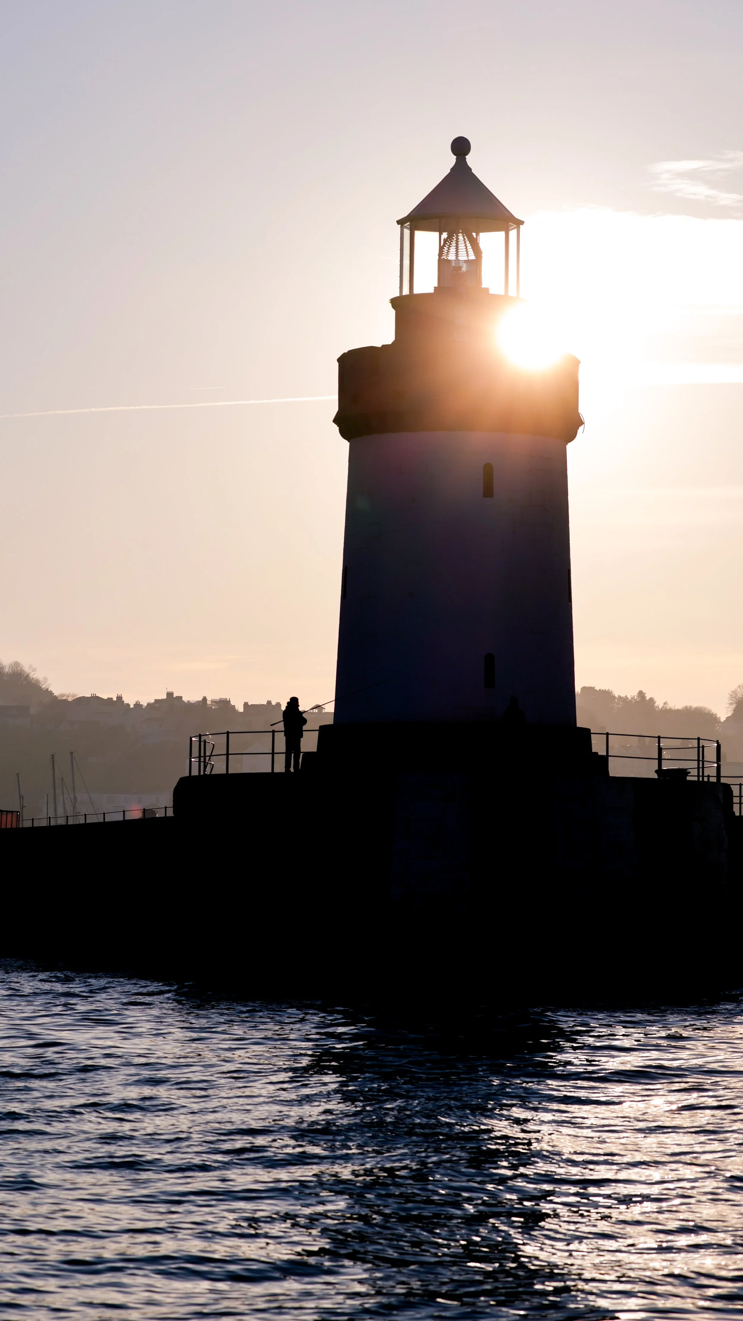 A lighthouse silhouetted against the setting sun, with a person fishing on the pier in the foreground, calm water in the front, and distant land and trees in the background.