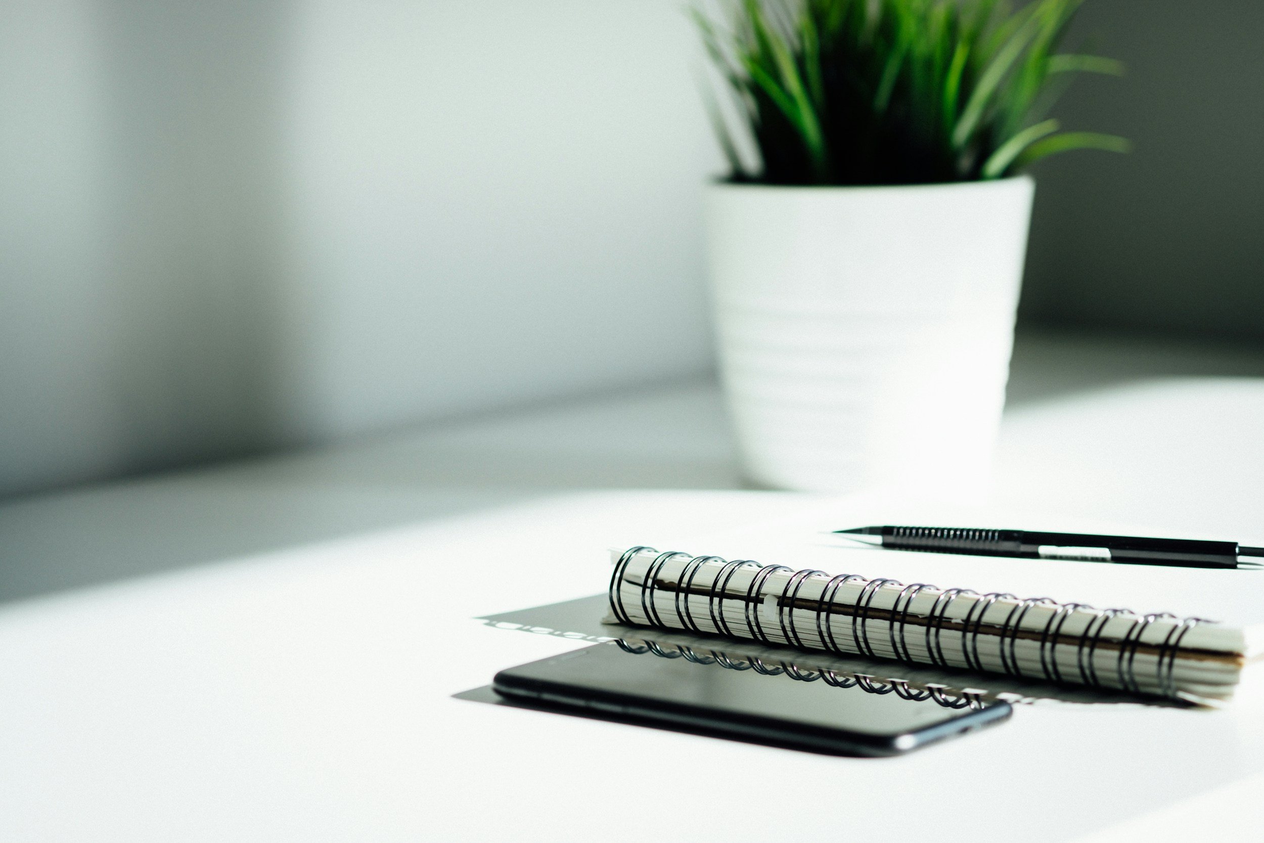 A white desk with a black notebook, a spiral-bound notepad, a pen, and a smartphone, with a white potted plant in the background.