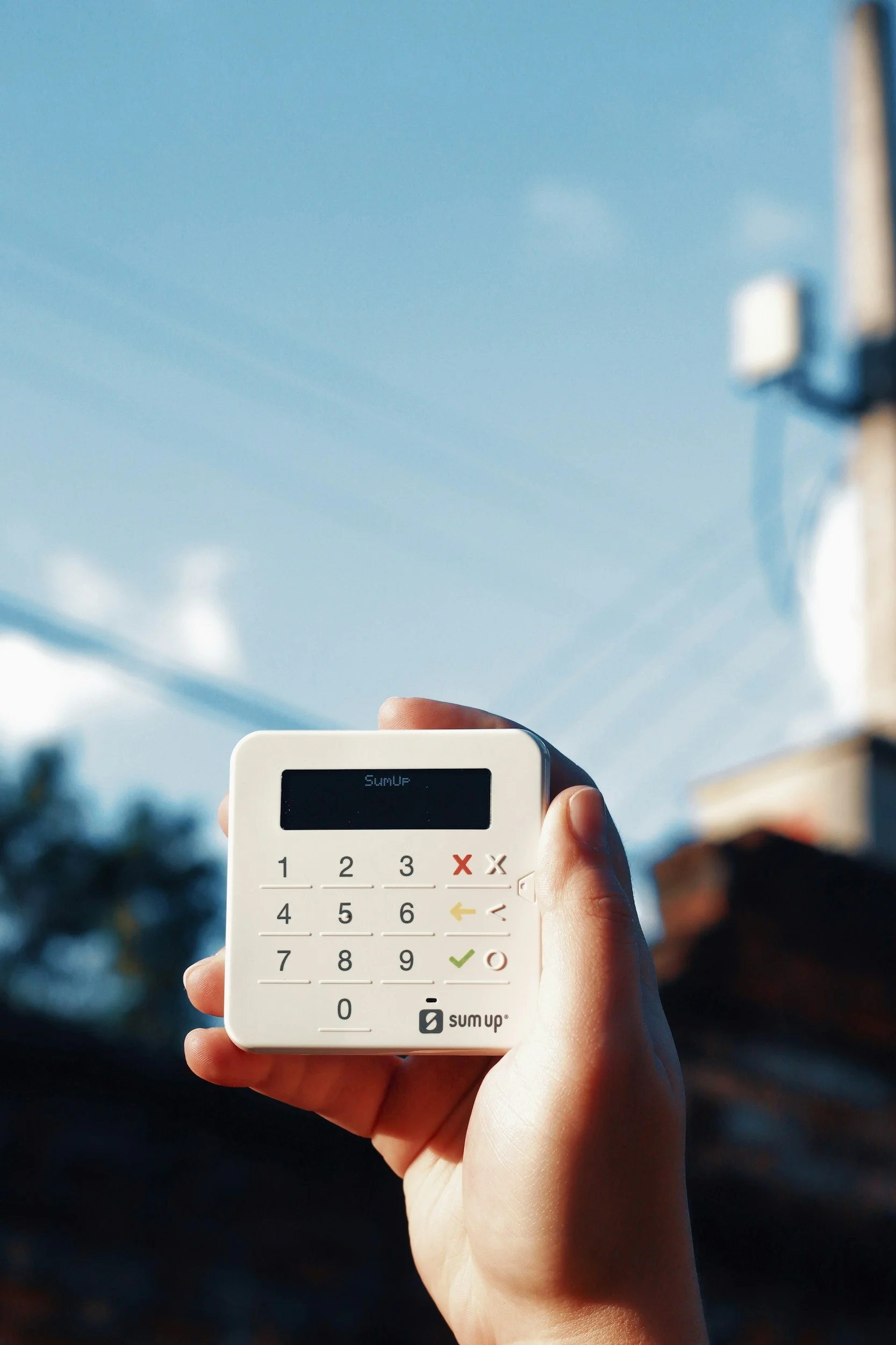 A hand holding a small white calculator with a digital display and colorful buttons, against a bright blue sky with some clouds.
