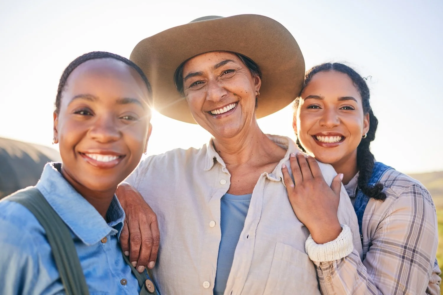 Three women smiling and embracing each other outdoors with the sun shining, one wearing a wide-brimmed hat.