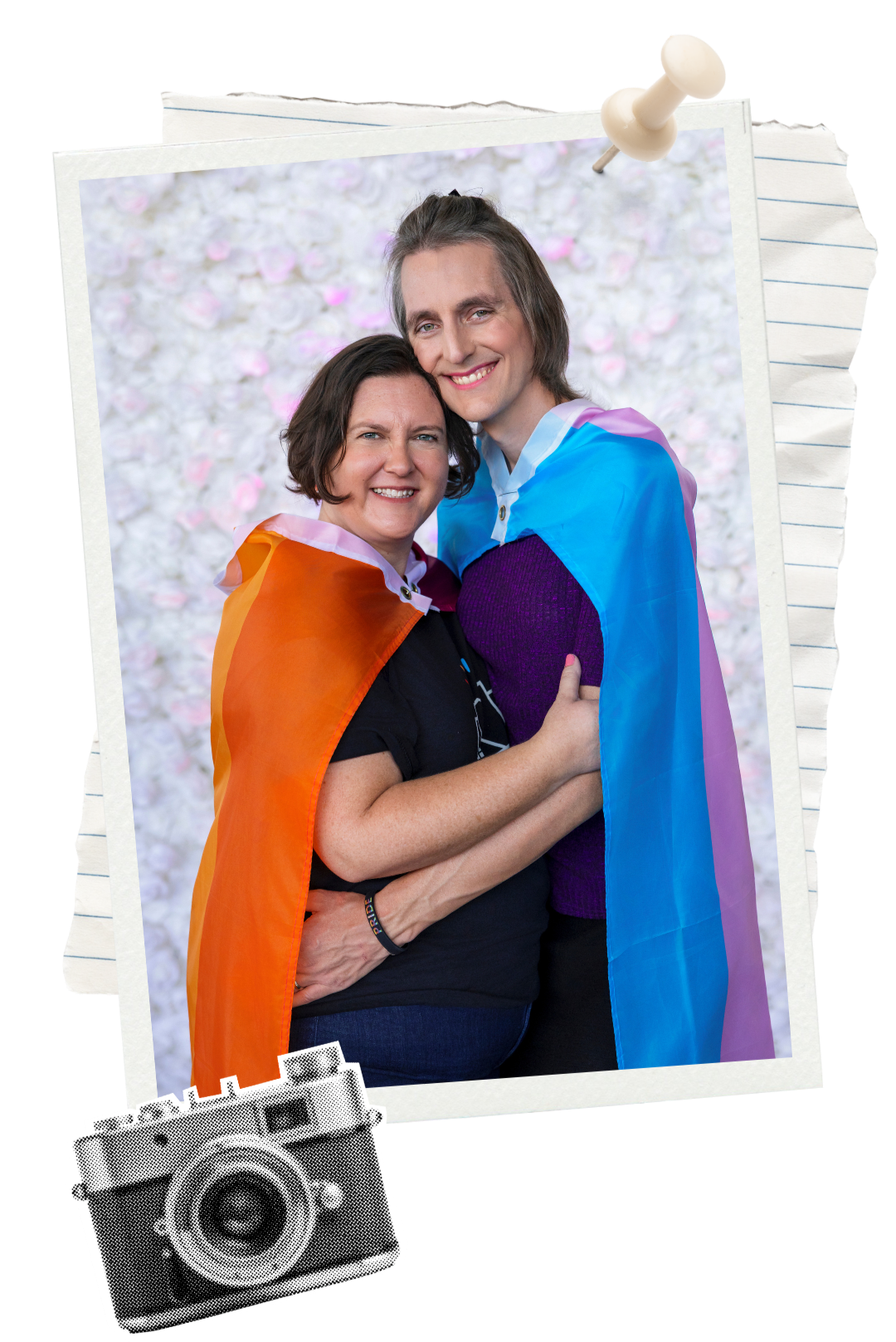 Amanda Pence and spouse smiling and embracing each other, wrapped in rainbow flags, standing against a background of pink and white flowers.