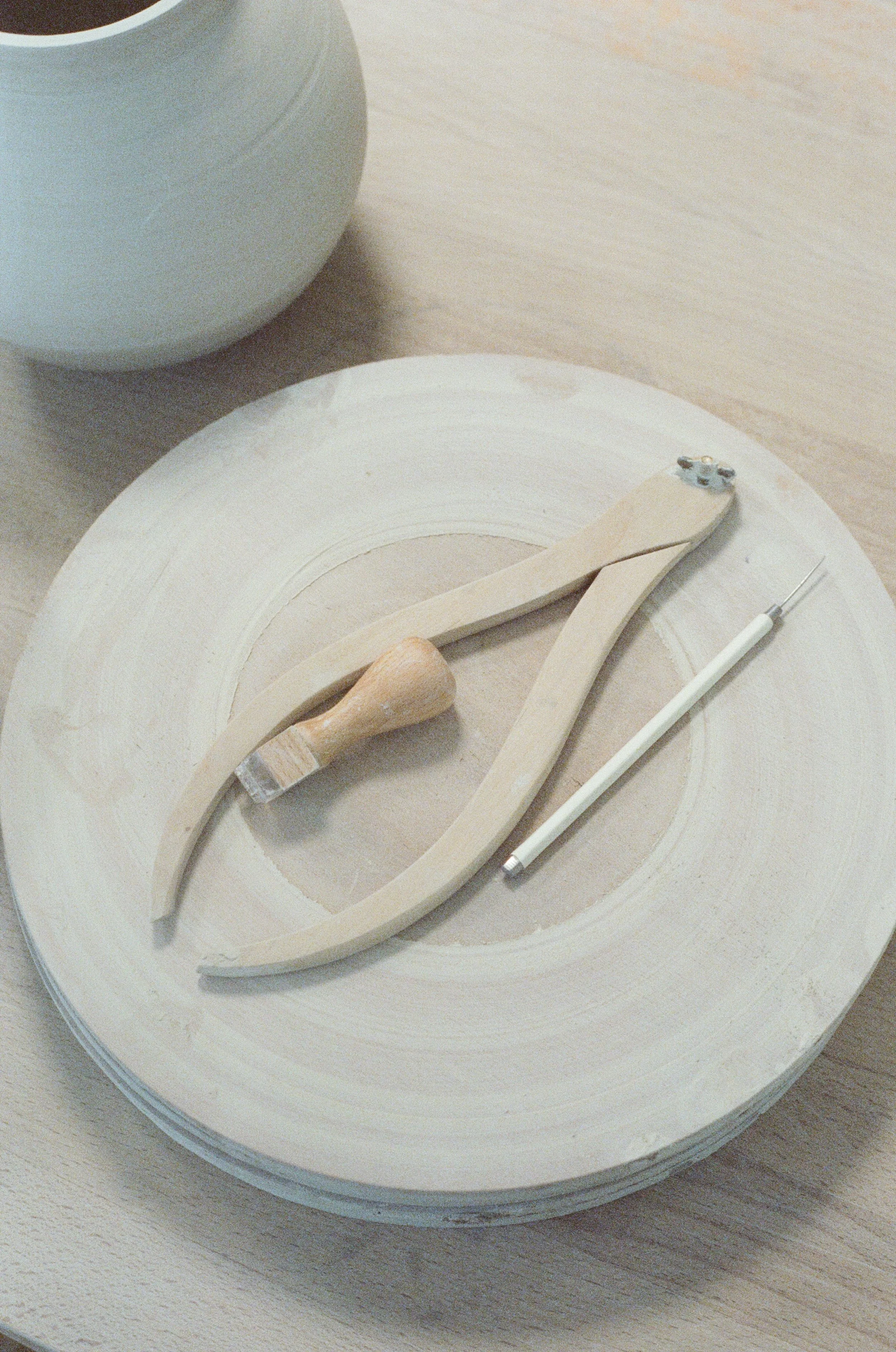 Un plateau avec plusieurs outils de poterie, y compris des rondeaux en bois, un pinceau, un compas, et un tampon, avec un bol blanc en porcelaine à côté.