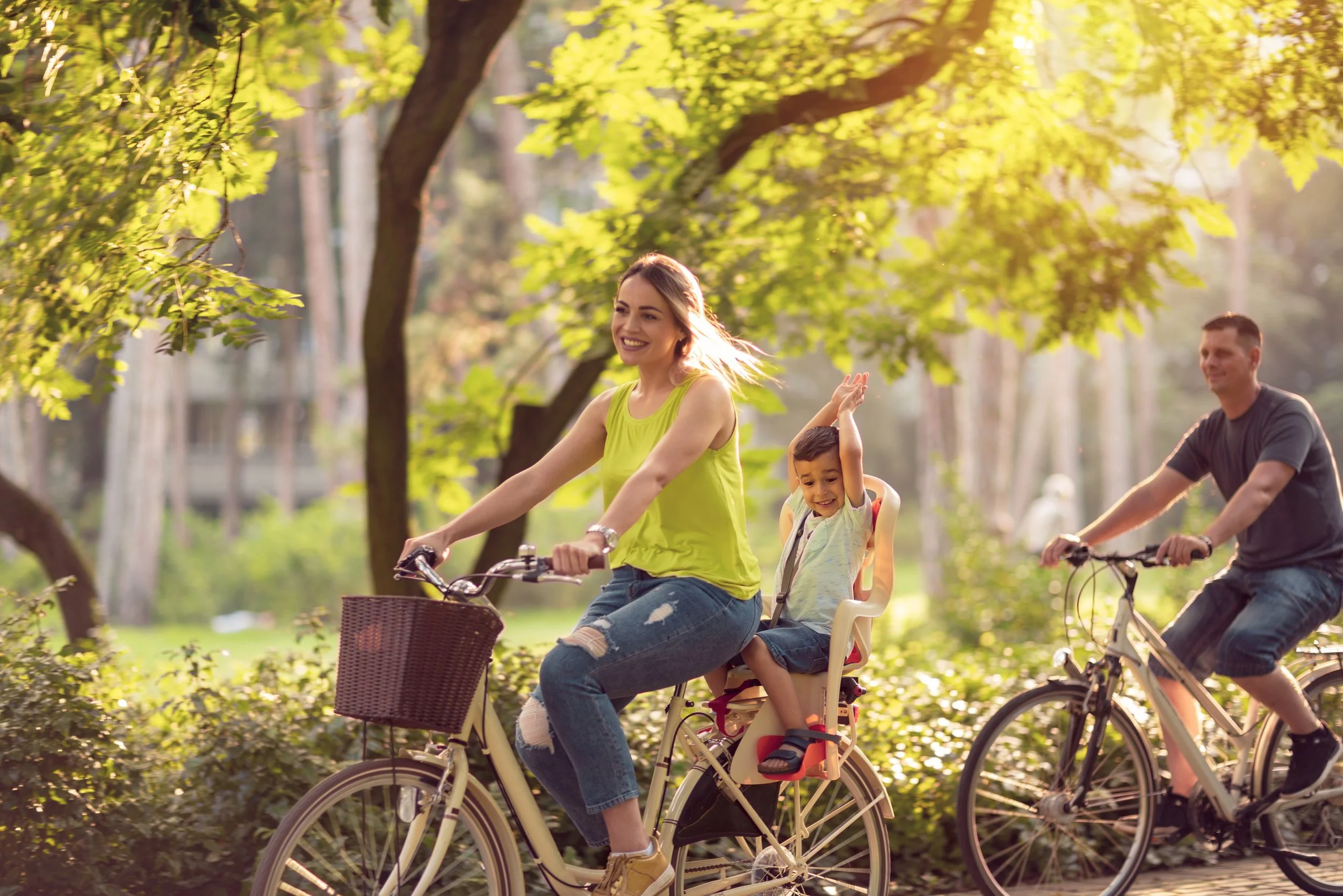 Young family riding bikes together along a shaded, tree-lined path on a sunny day