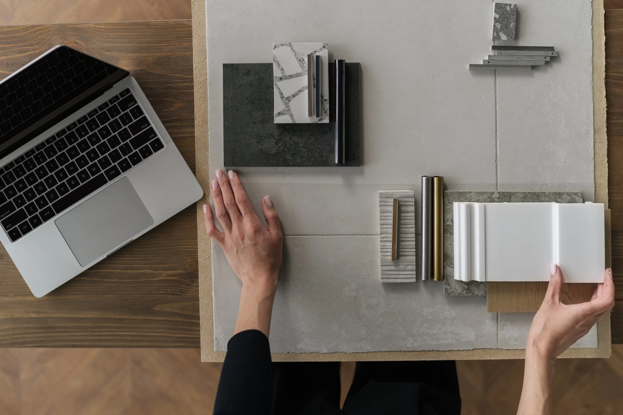Hands arranging tile, countertop, trim and cabinet hardware samples on a table beside a laptop