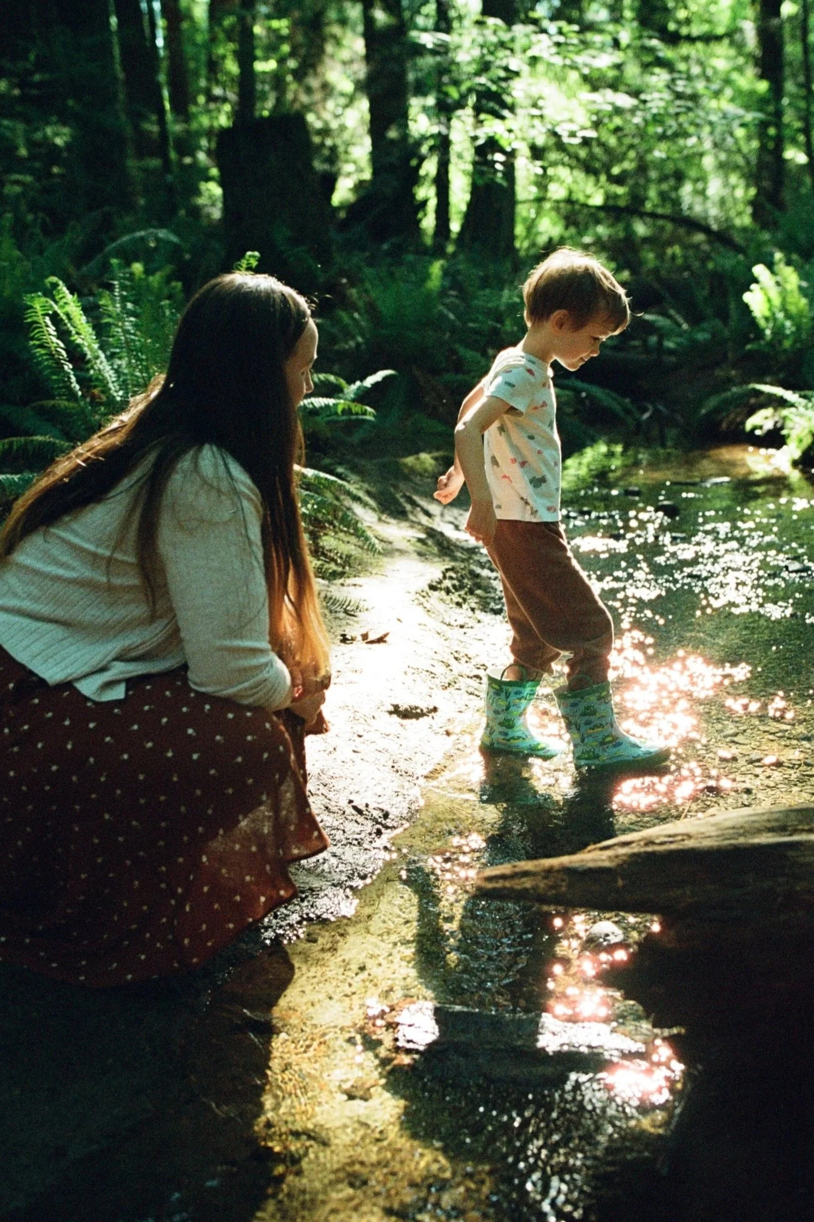 A woman crouches by a stream, watching her kiddo wading happily in the water, wearing rubber boots. Shot by Seattle photographer Holly Stevens.