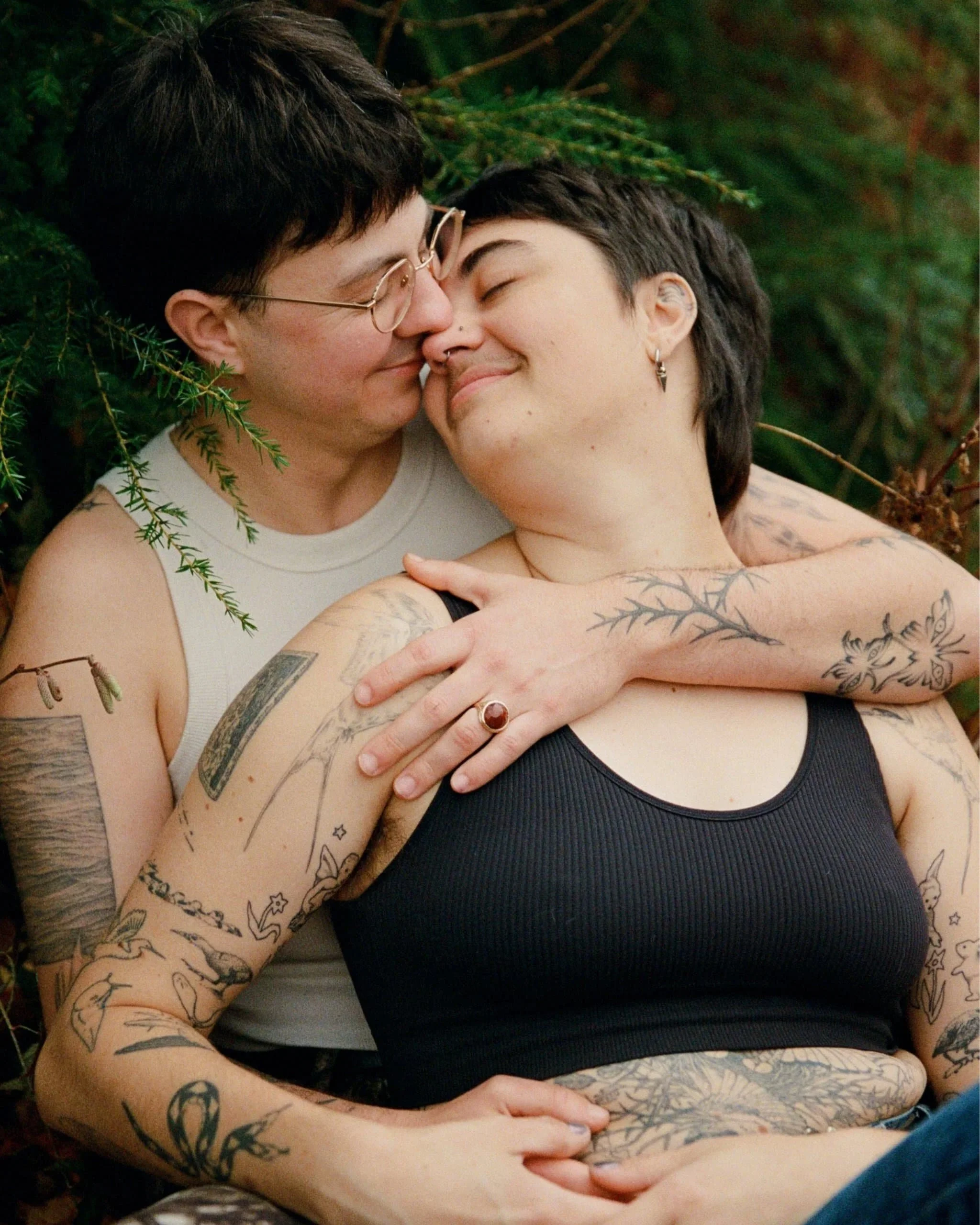 Two individuals with short dark hair and tattoos, hugging outdoors among green foliage, smiling and appearing affectionate. Shot by photographer Holly Stevens.