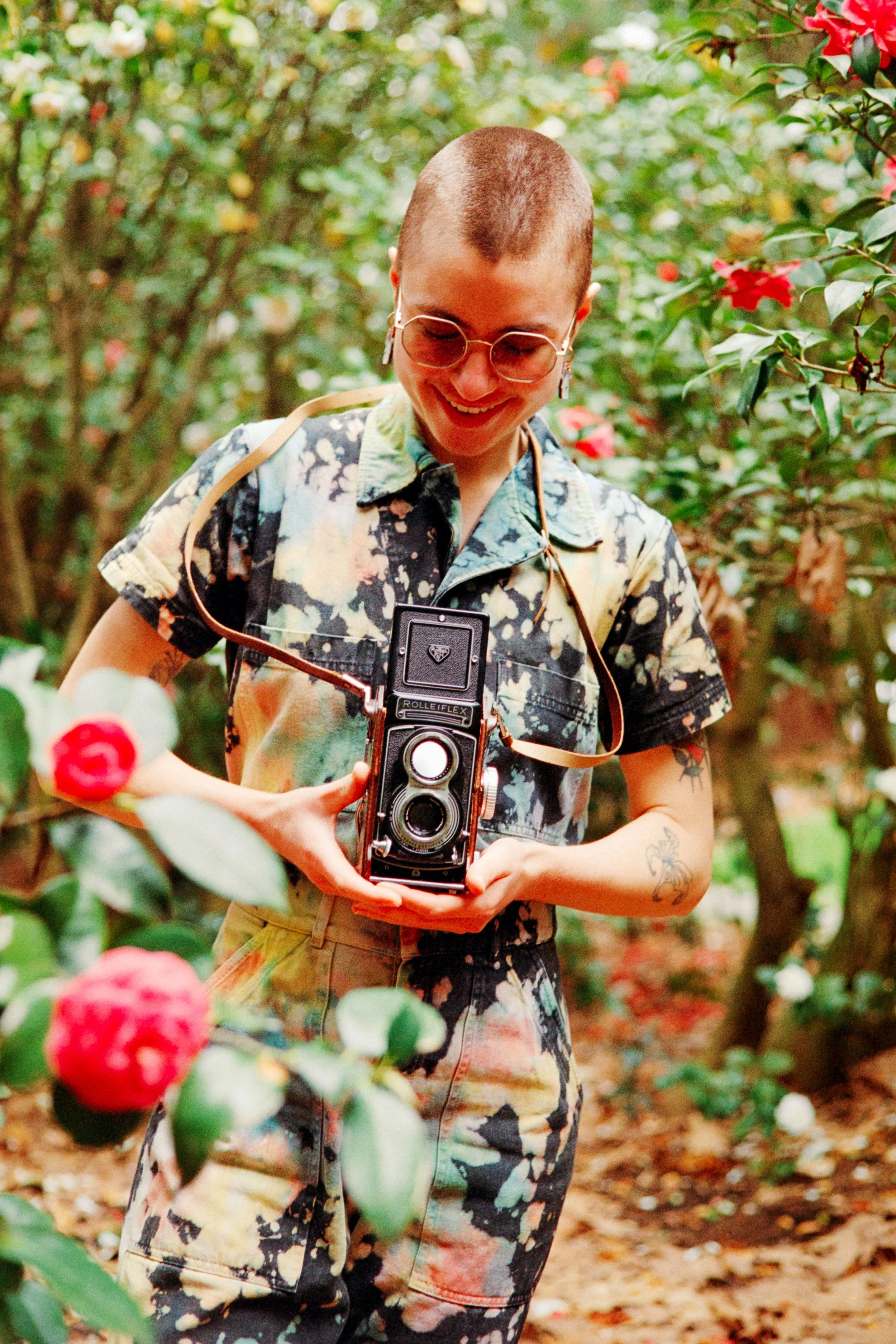 Person with a buzz cut hairstyle, wearing textured glasses, floral patterned shirt and cargo pants, holding a vintage camera in a garden with flowers and greenery.