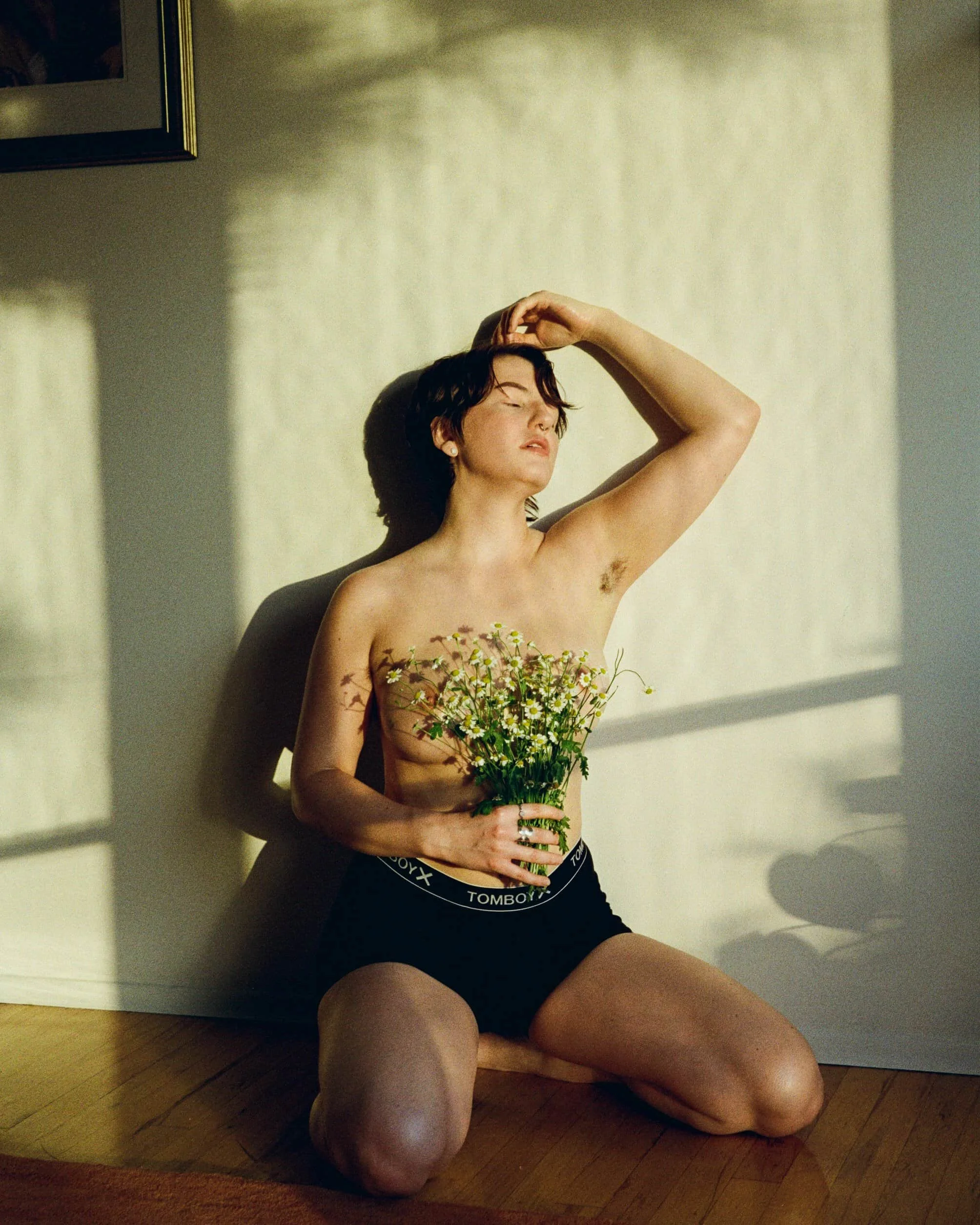 A nonbinary person in Seattle with short dark hair kneels on a hardwood floor, holding a bouquet of small white flowers, with the sunlight casting shadows on the wall behind them.