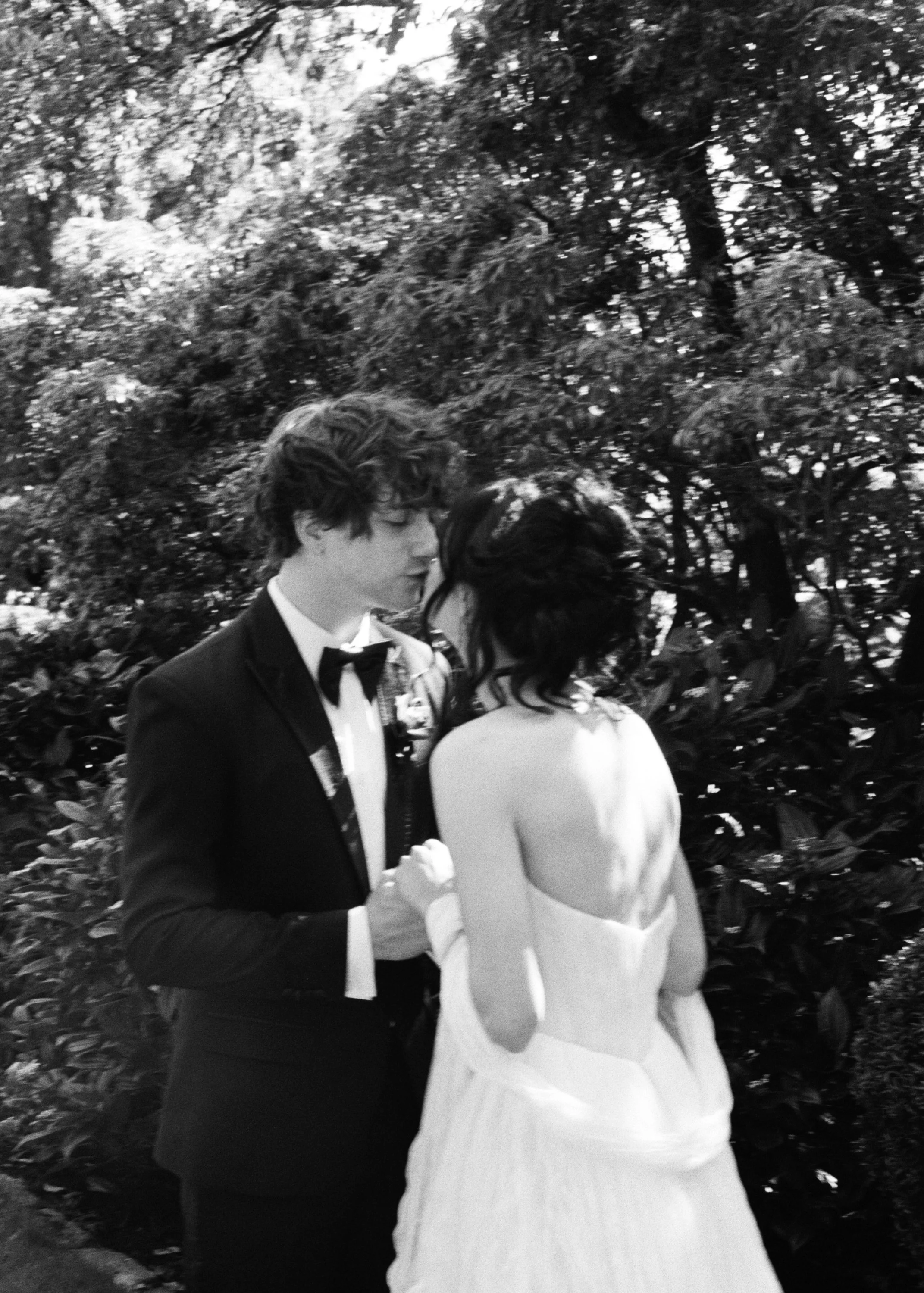 A nonbinary person in a tuxedo kisses a dark-haired woman in a wedding dress at Parsons Gardens. Photographed by queer photographer Holly Stevens.