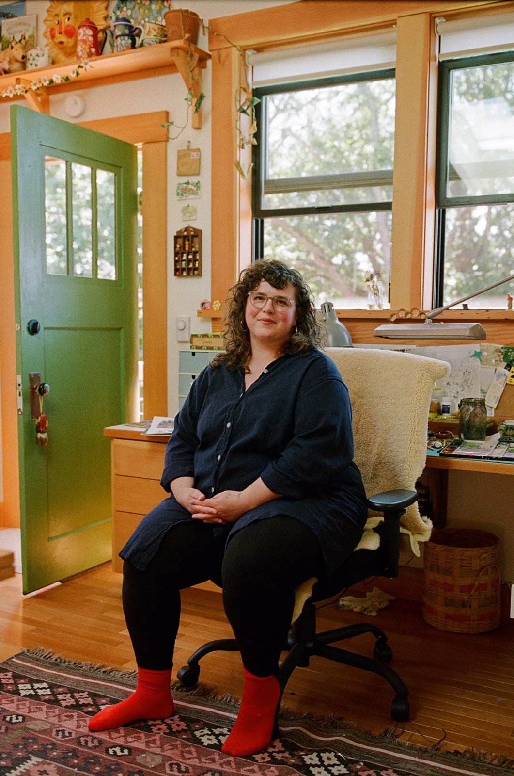 Illustrator Phoebe Wahl sits in her studio with her hands clasped, smiling softly into the camera. Shot by Seattle photographer Holly Stevens.