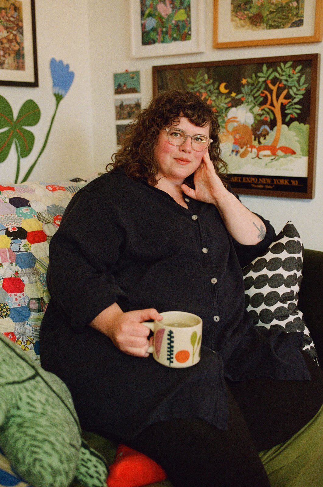 A woman with curly brown hair and glasses sitting on a couch, holding a mug with a colorful pattern, surrounded by framed artwork and colorful cushions. Shot by photographer Holly Stevens.