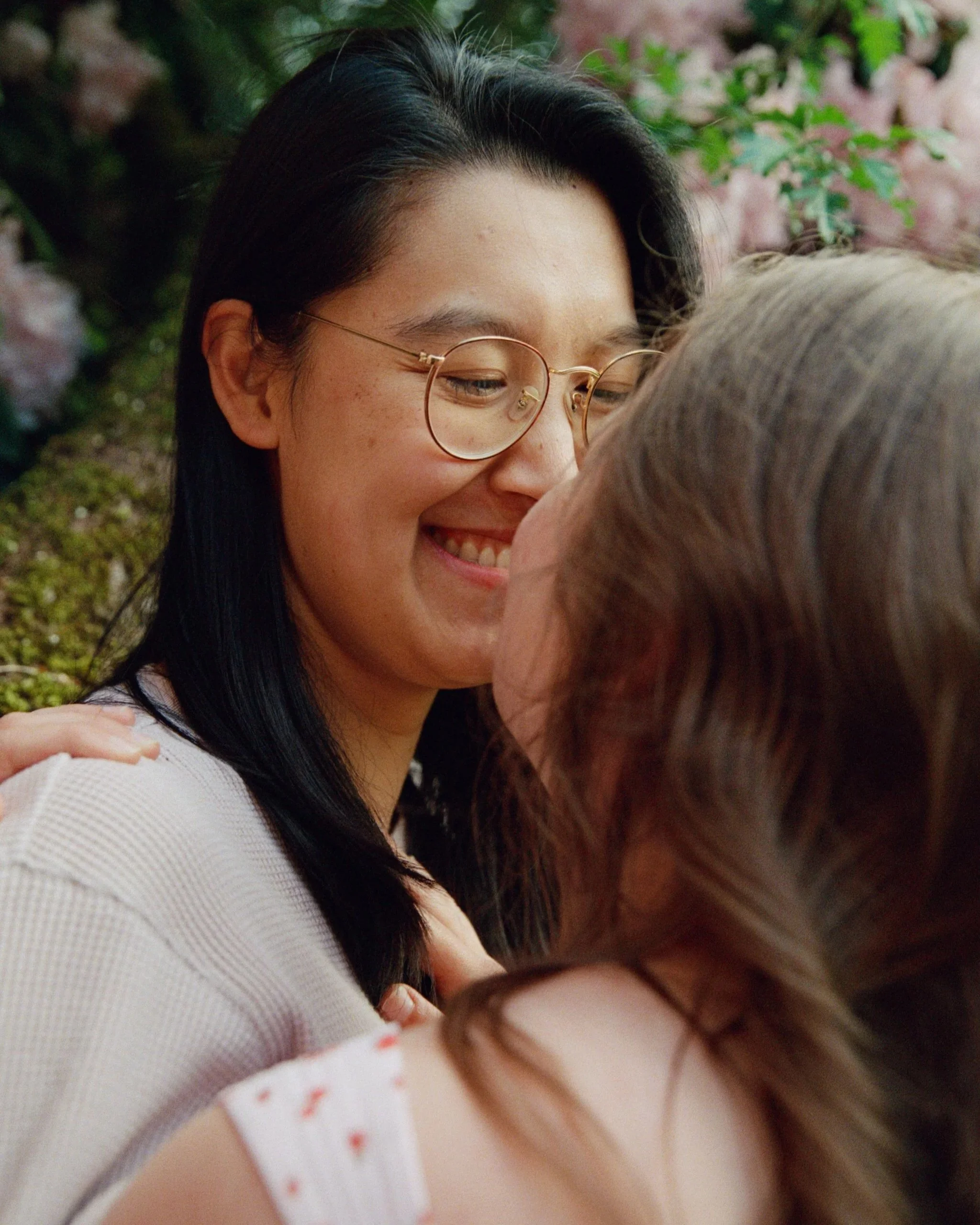 A young woman with glasses and black hair smiling as she embraces an elderly woman in a garden with pink flowers.