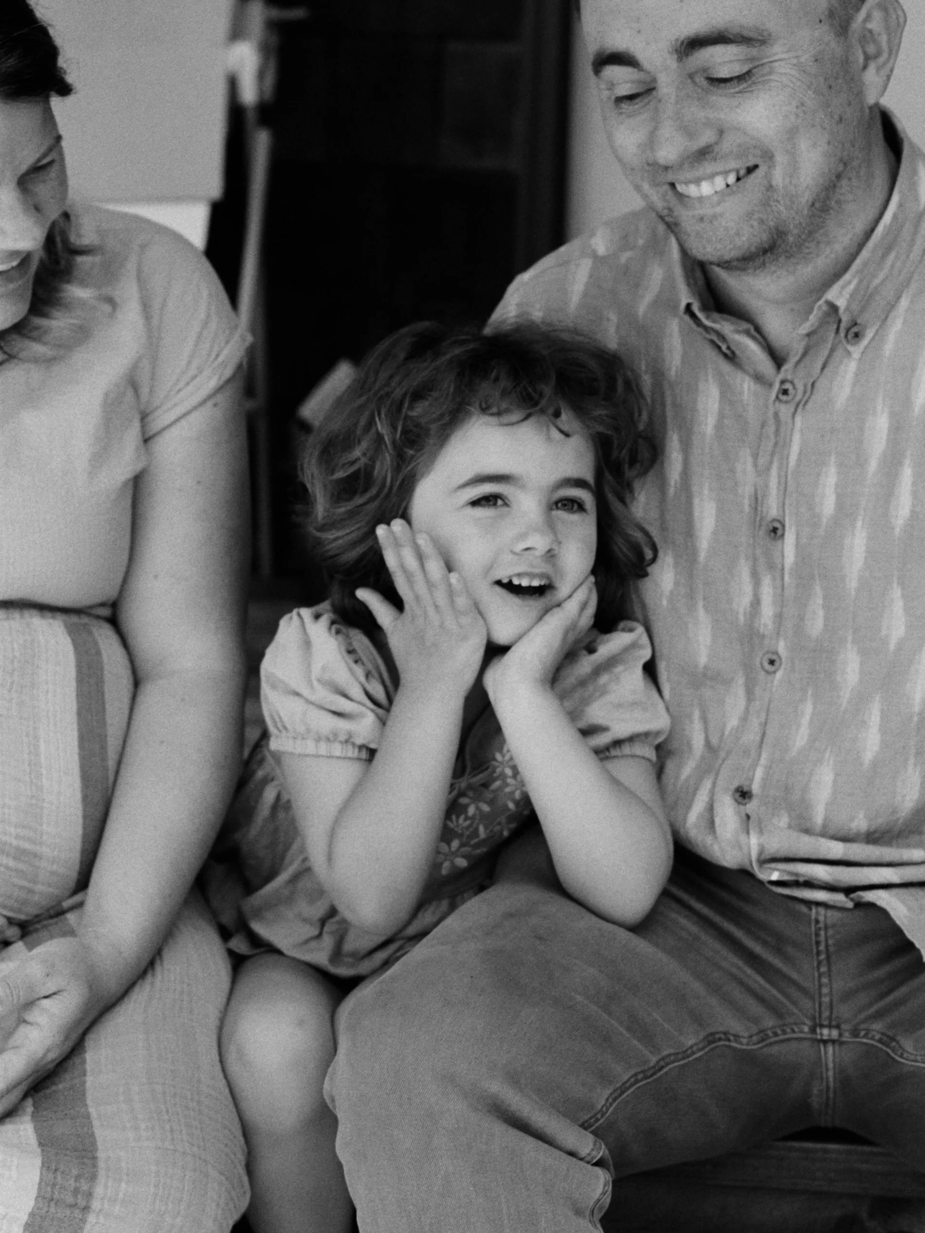 A black and white photo of a family scene showing a woman, man, and young girl. The girl is sitting on the man's lap, holding her face with her hands, smiling and looking towards the camera. The man is smiling and looking down at the girl. The woman is partially visible, looking towards the girl. The scene appears warm and affectionate.