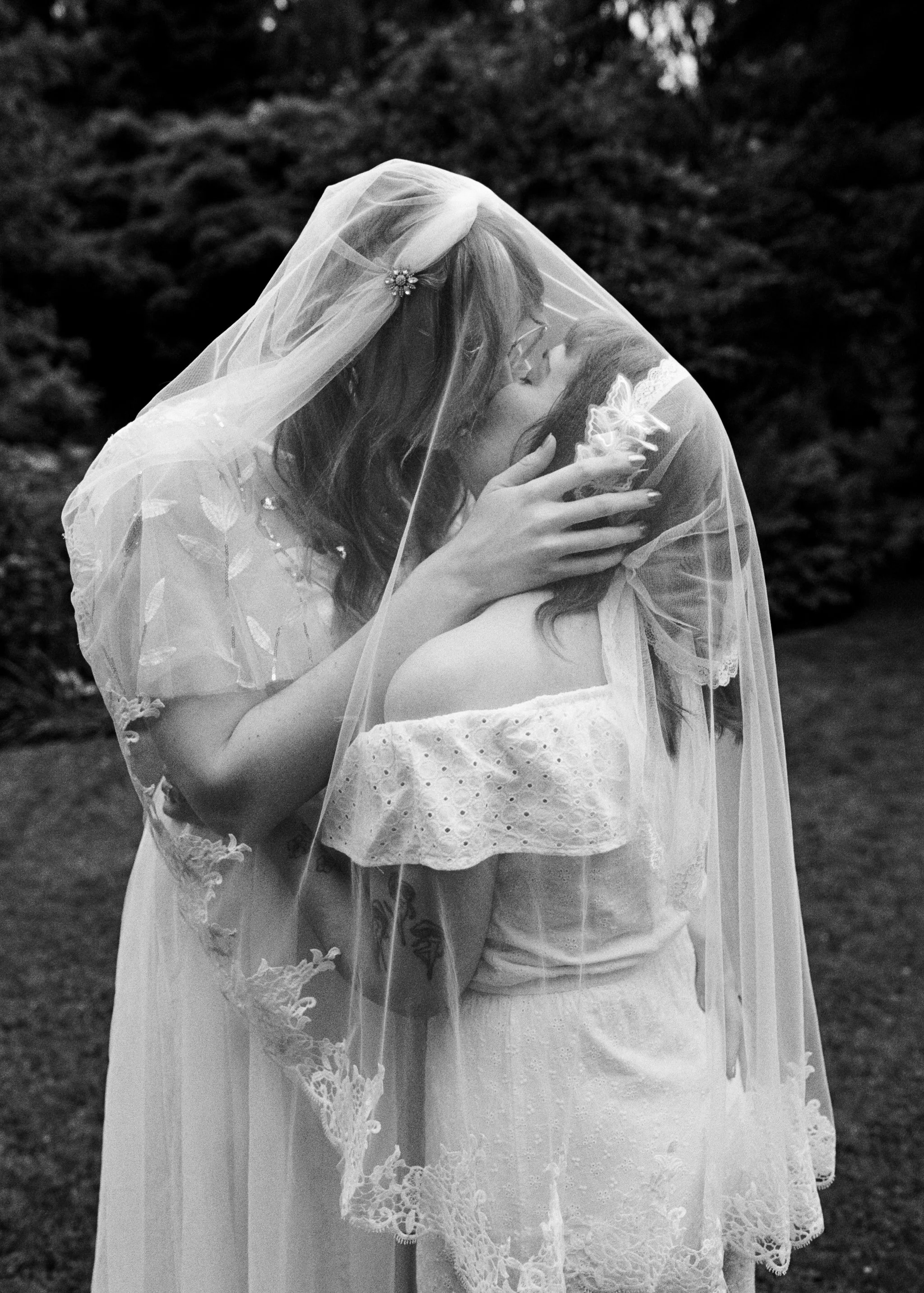Two women in wedding dresses sharing an intimate moment, with one woman wearing a veil.