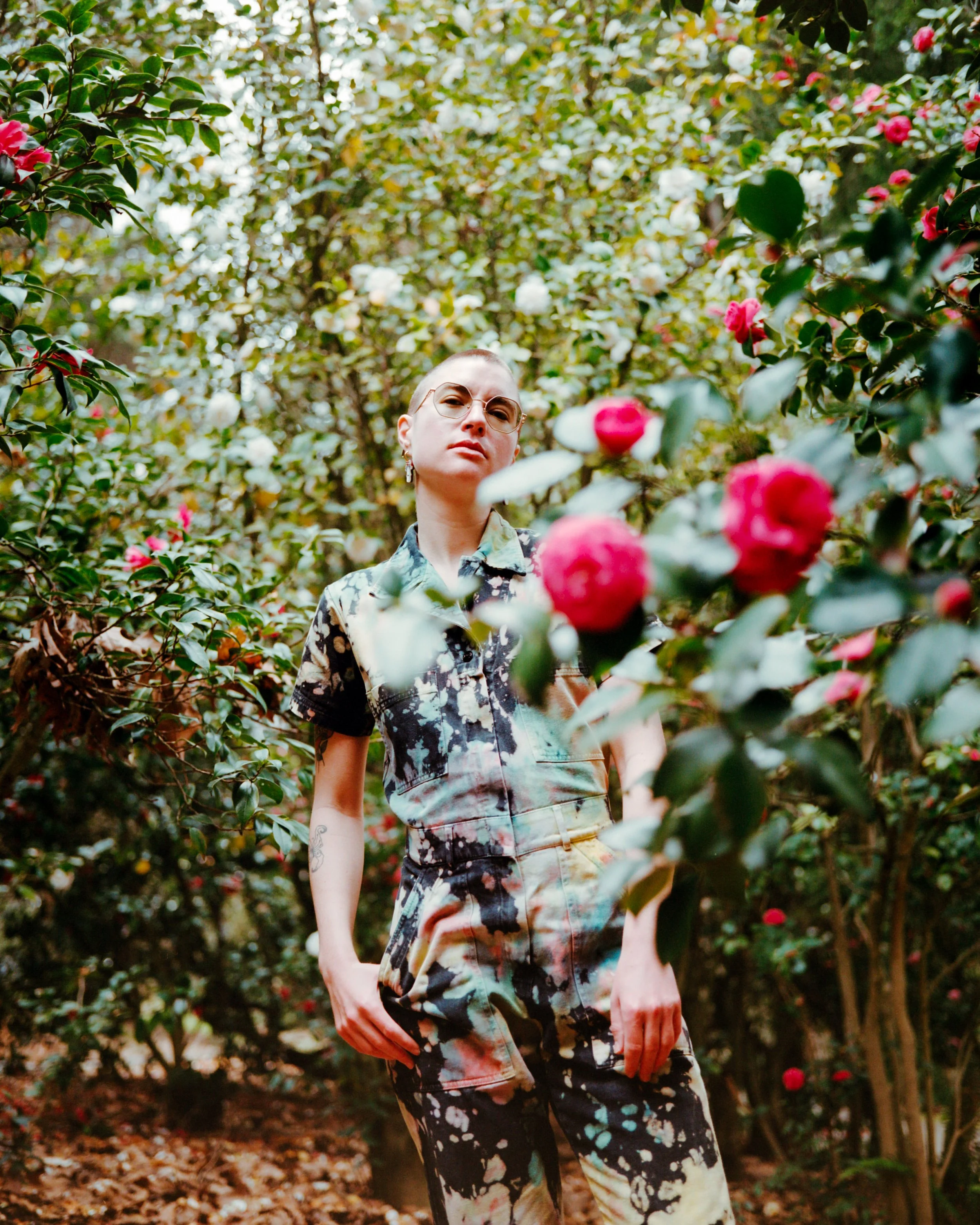 A person with short hair, glasses, and earrings stands amidst dense shrubbery with pink and white flowers, wearing an artistic, colorful outfit.