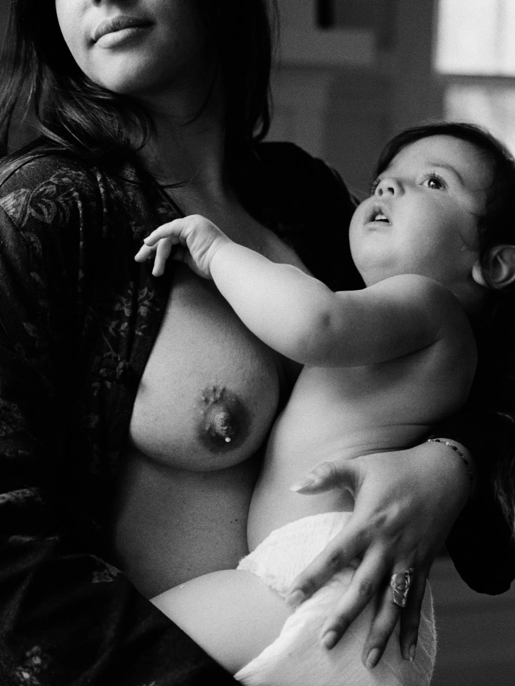 A queer woman in Seattle holding a young child, who is reaching towards her breast, in a black-and-white photograph.