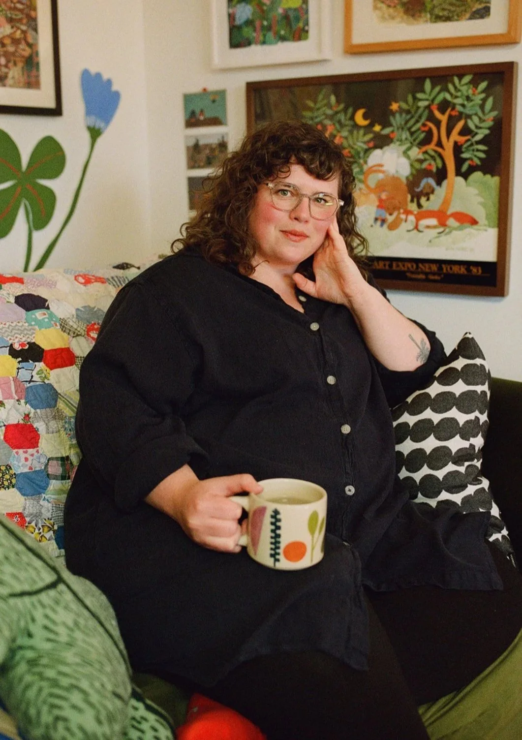 A woman with curly brown hair and glasses sitting on a couch, holding a mug with a colorful pattern, surrounded by framed artwork and colorful cushions. Shot by Seattle-based queer photographer Holly Stevens.