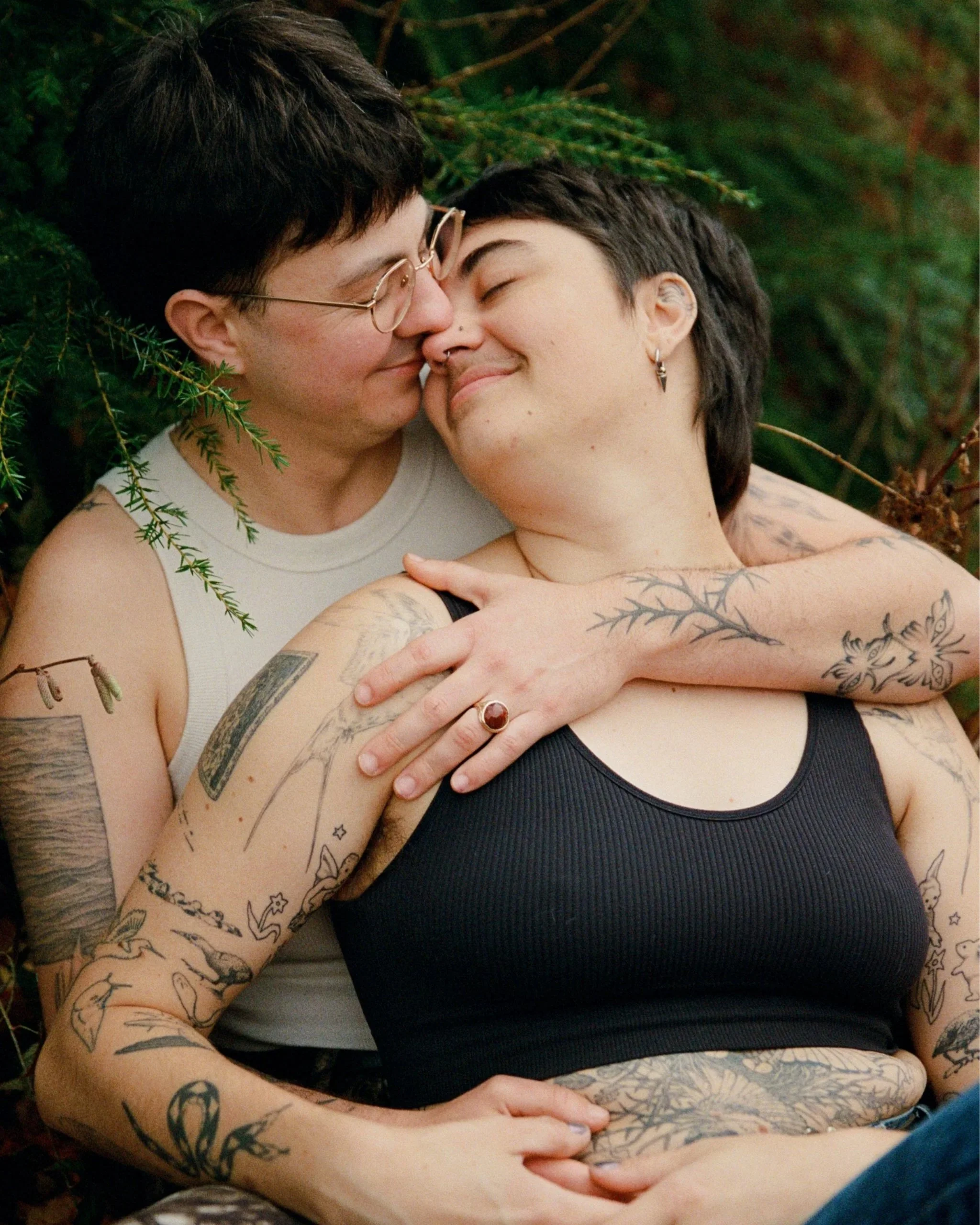 Two women with tattoos embracing outdoors, with one wearing glasses and a white tank top, and the other in a black tank top. They are smiling, with their foreheads touching amid green foliage.