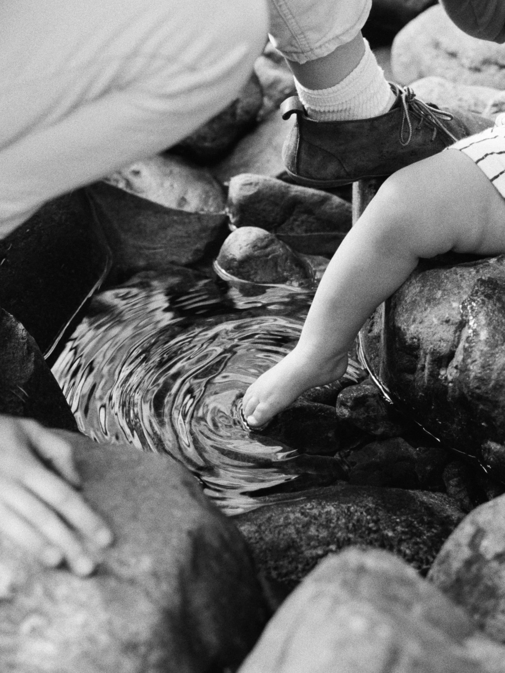 Black and white photo of a child dipping their bare foot into a small pool of water surrounded by rocks, with an adult’s hand and leg nearby.