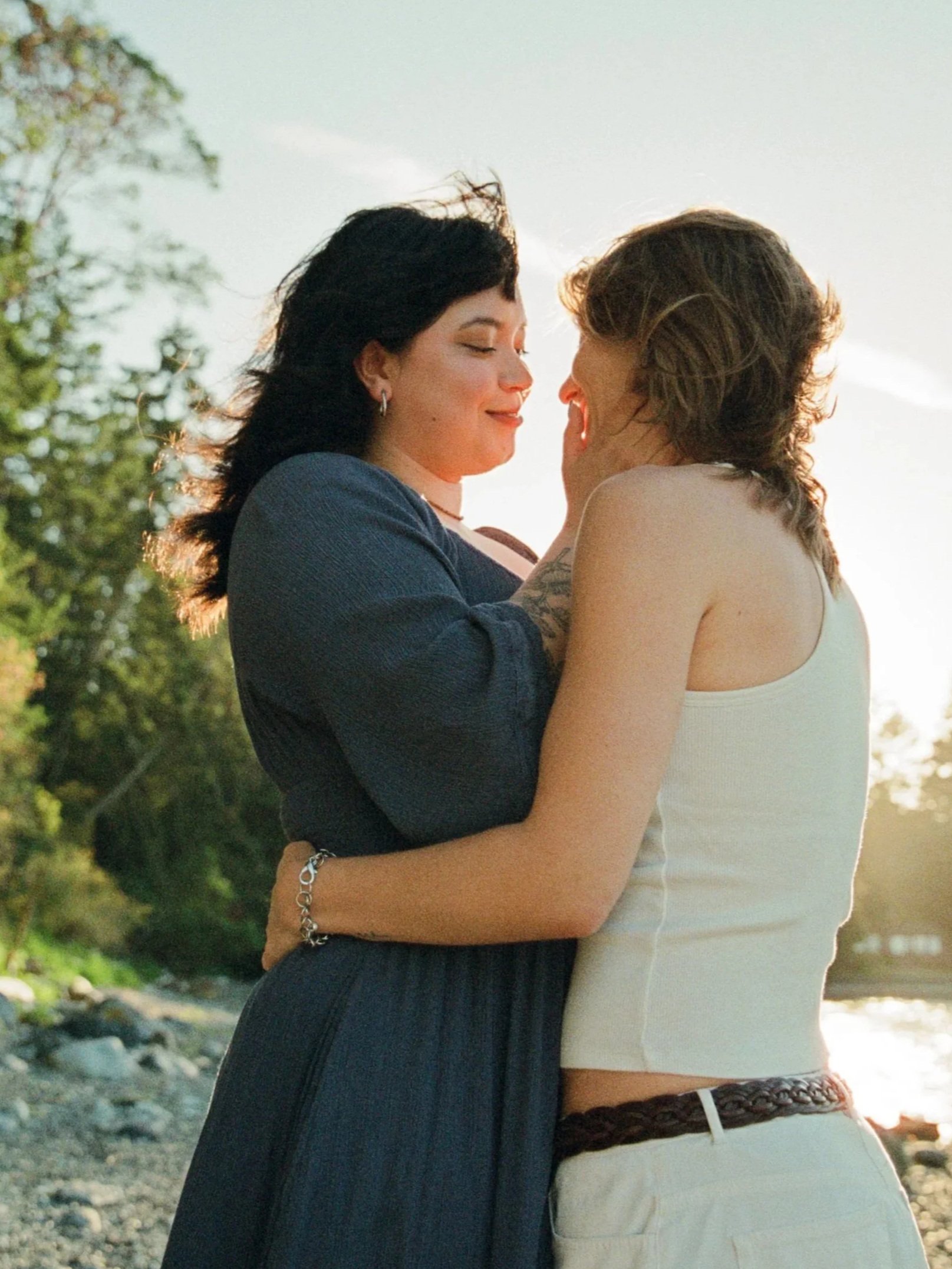 A couple standing close together outdoors during sunset, smiling and gazing into each other's eyes, with trees and a rocky ground in the background.
