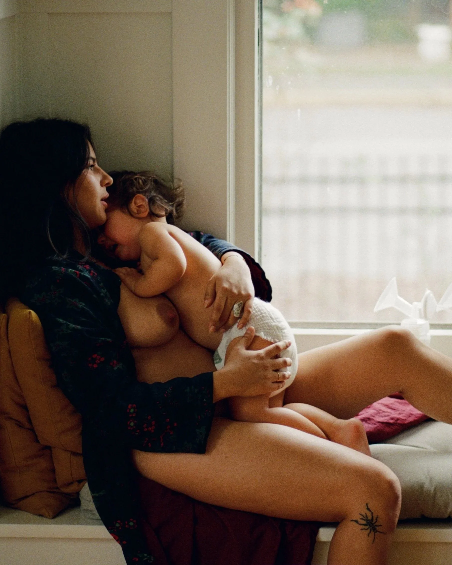 A woman with dark hair breastfeeding a young child on a couch in front of a window.