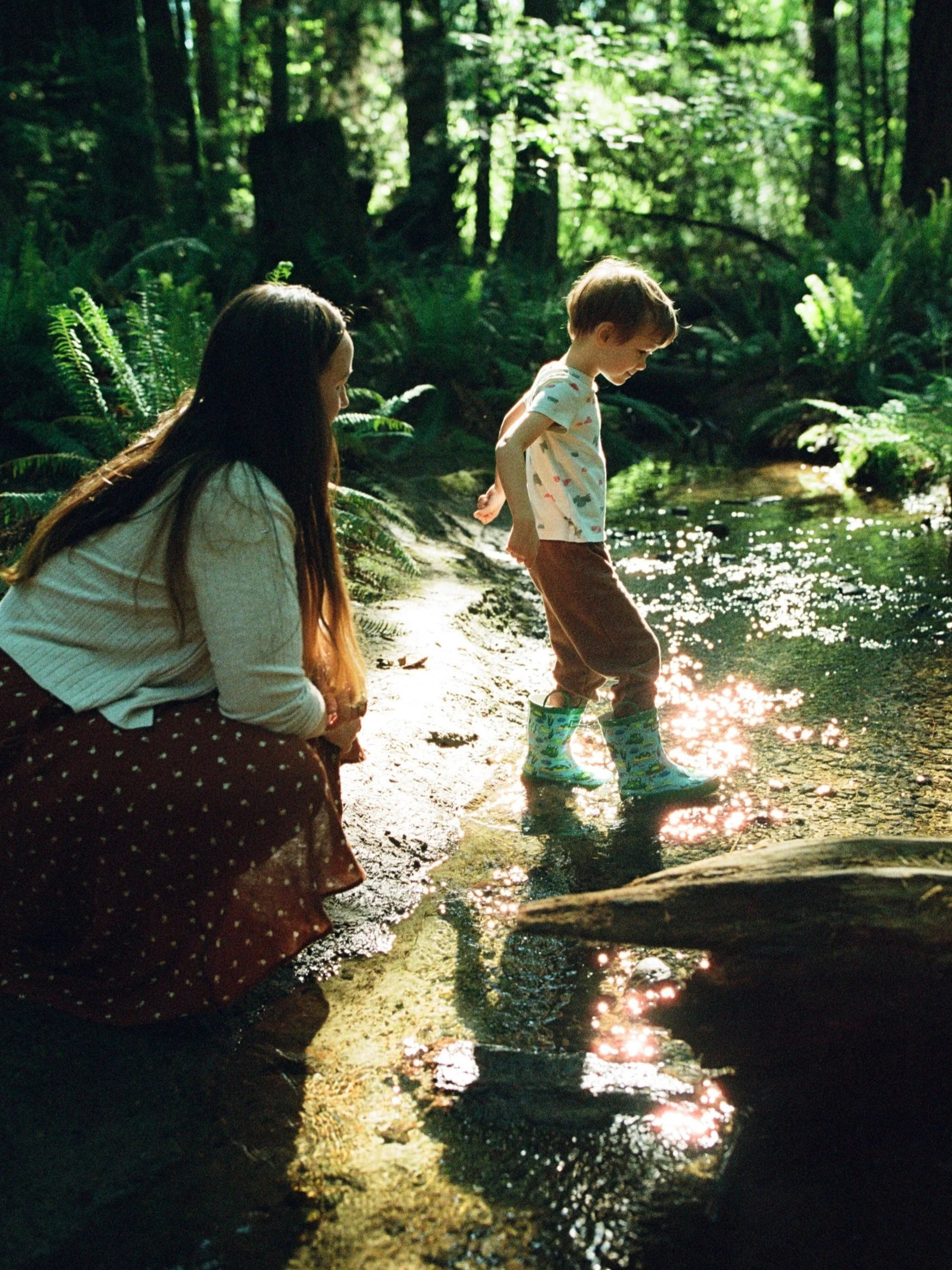 A woman with long brown hair kneels down next to a creek, where a young boy is wading in his rain boots. They both smile happily. Shot by queer photographer Holly Stevens.