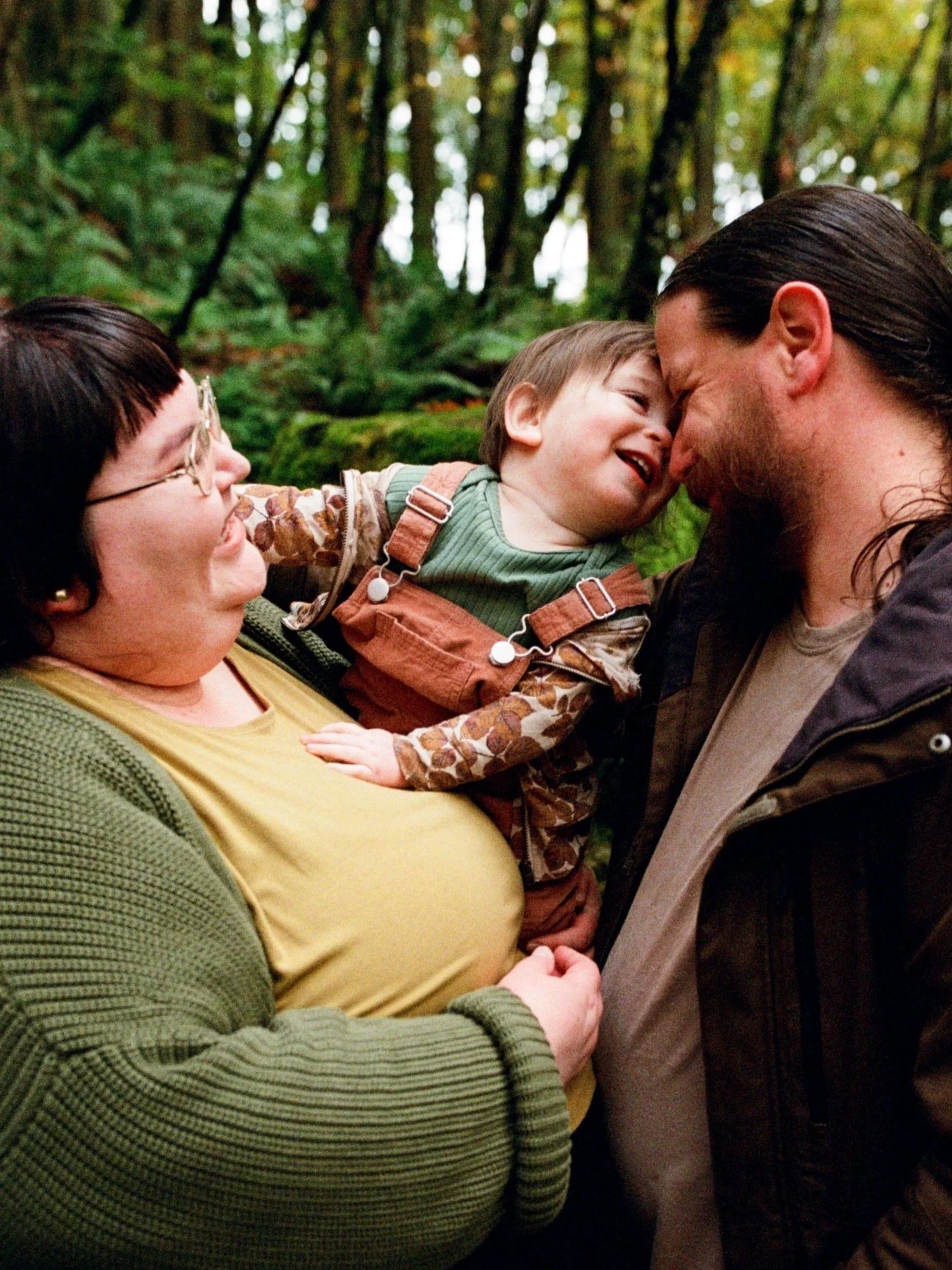 A smiling woman holds a young child who is touching noses and laughing with a bearded man in a forest, surrounded by green trees and ferns. Shot by Seattle photographer Holly Stevens.