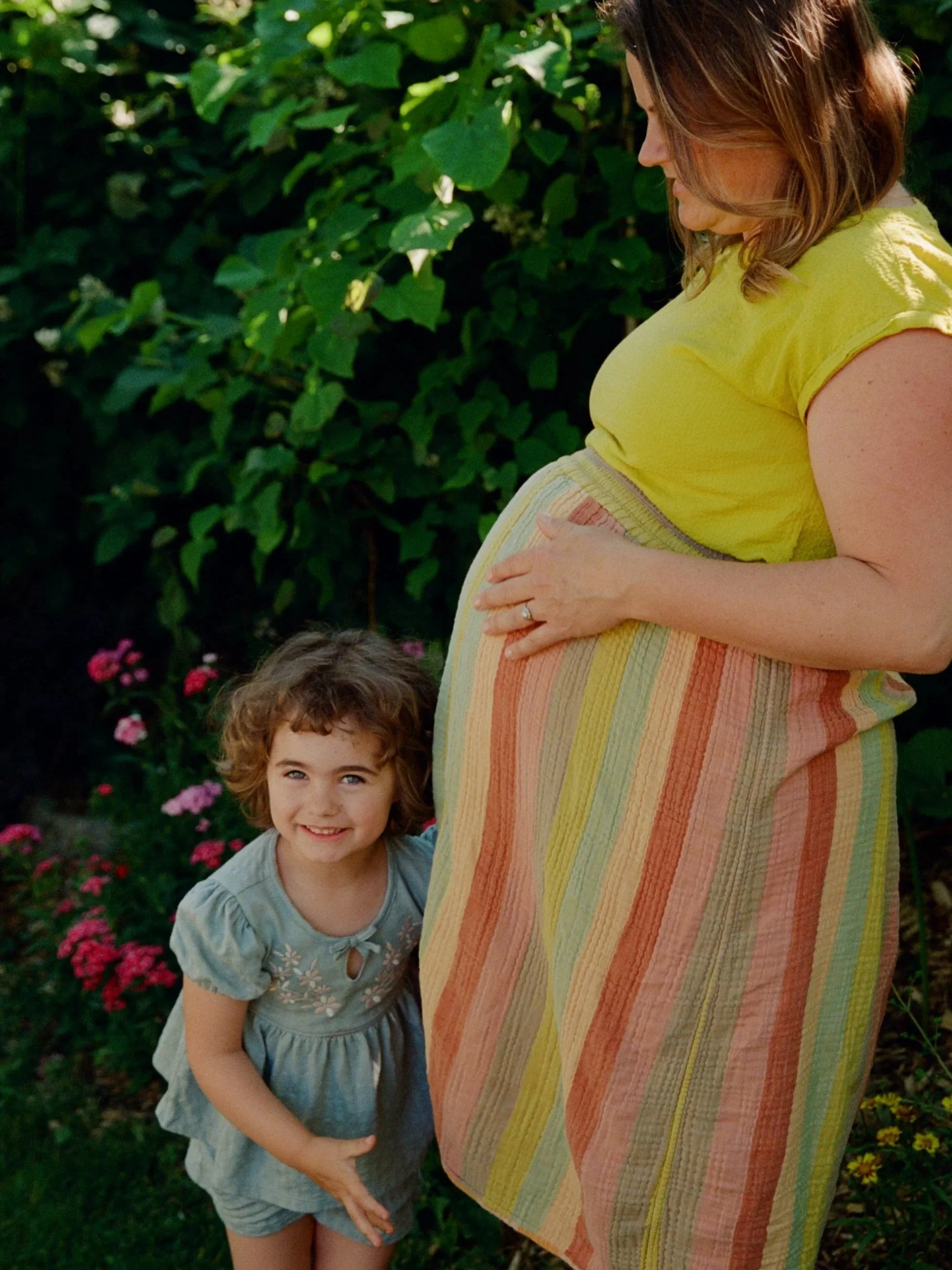 A pregnant woman in a yellow top and rainbow-colored skirt standing in a garden with green foliage, accompanied by a young girl with curly hair and a gray dress, smiling at the camera. Shot by queer photographer Holly Stevens.