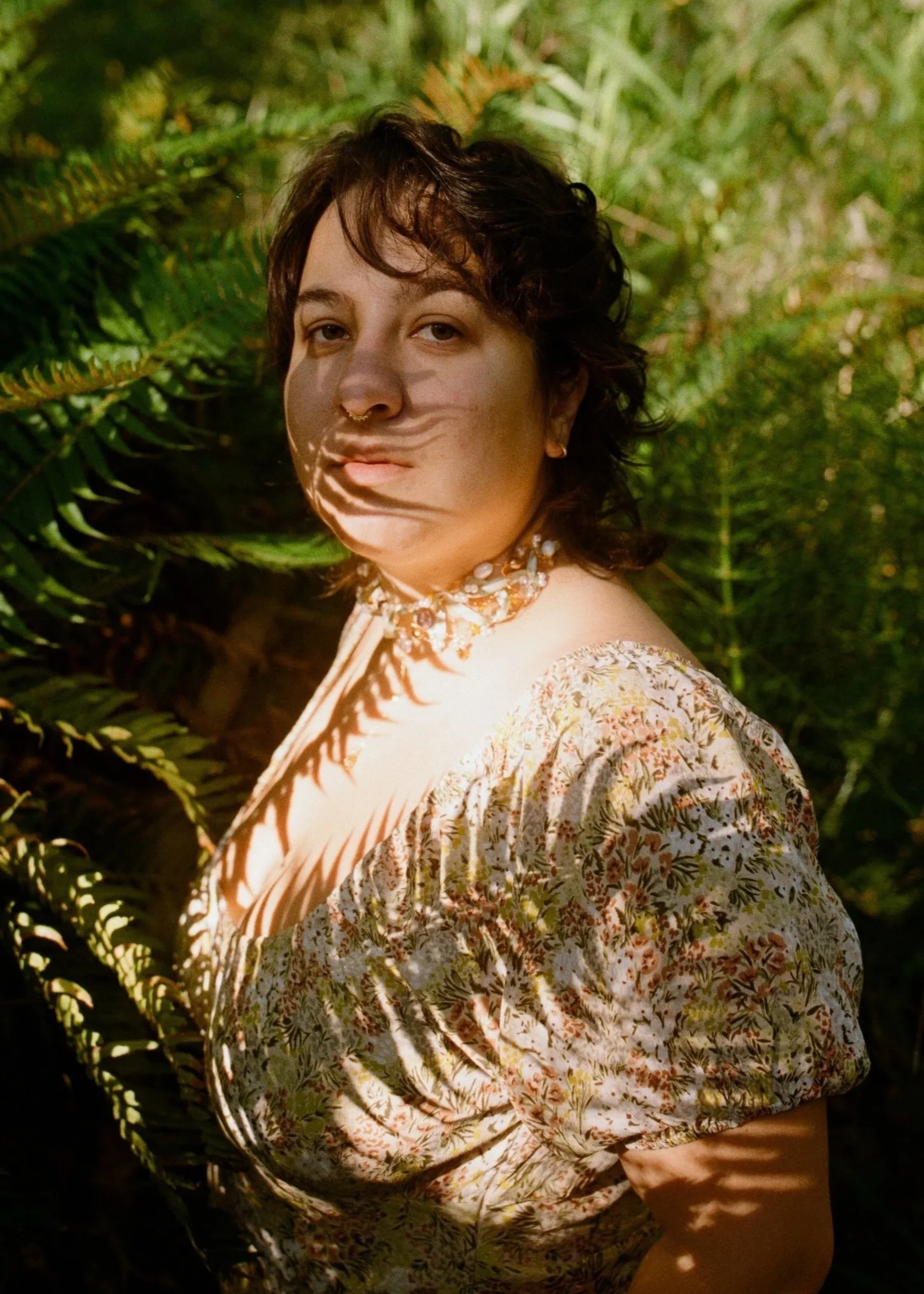 A woman with short curly hair and wearing a patterned dress and jewelry stands among green ferns, with sunlight creating shadows on her face.
