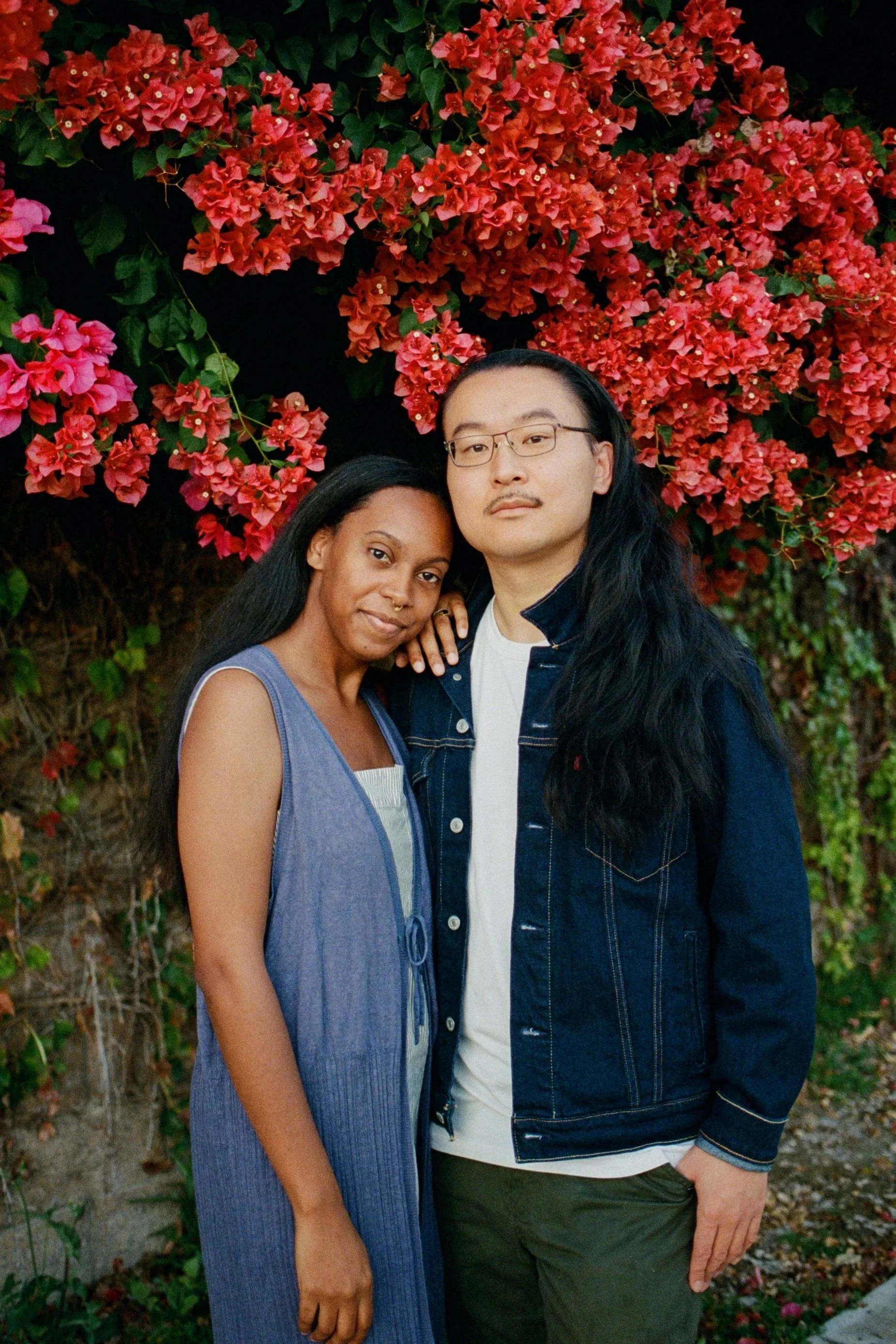 A woman with dark skin and long black hair smiling, standing close to a man with long black hair, glasses, and a slight mustache, in front of a large bush with pink and red flowers. The woman is wearing a sleeveless purple dress, and the man is wearing a black jacket over a white shirt and dark green pants.