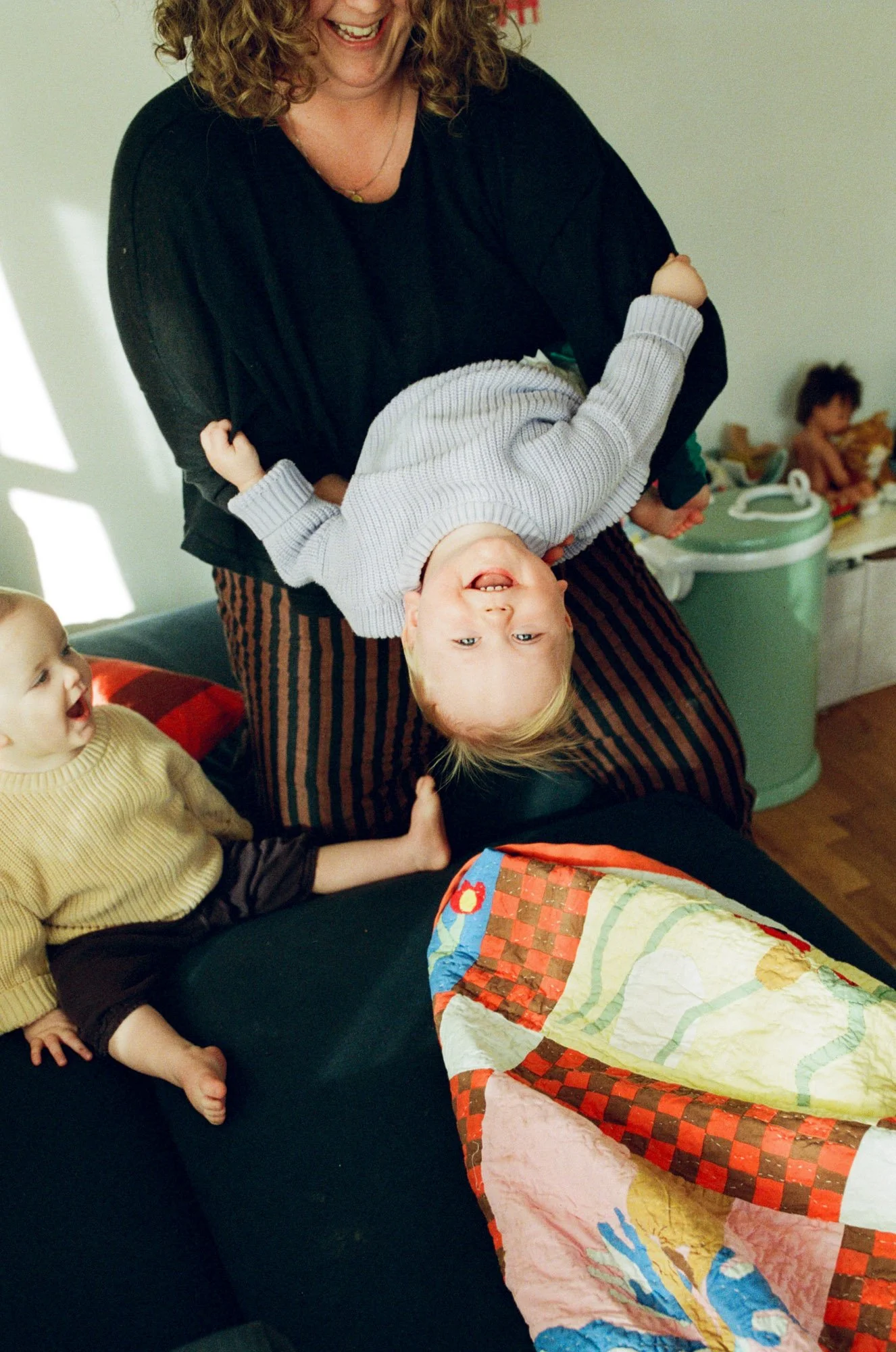 A woman holding a young boy upside down, both smiling and enjoying the moment. A toddler sitting on a couch nearby is looking at them, and a quilt is draped over an object in the foreground. Shot by Seattle portrait photographer Holly Stevens.