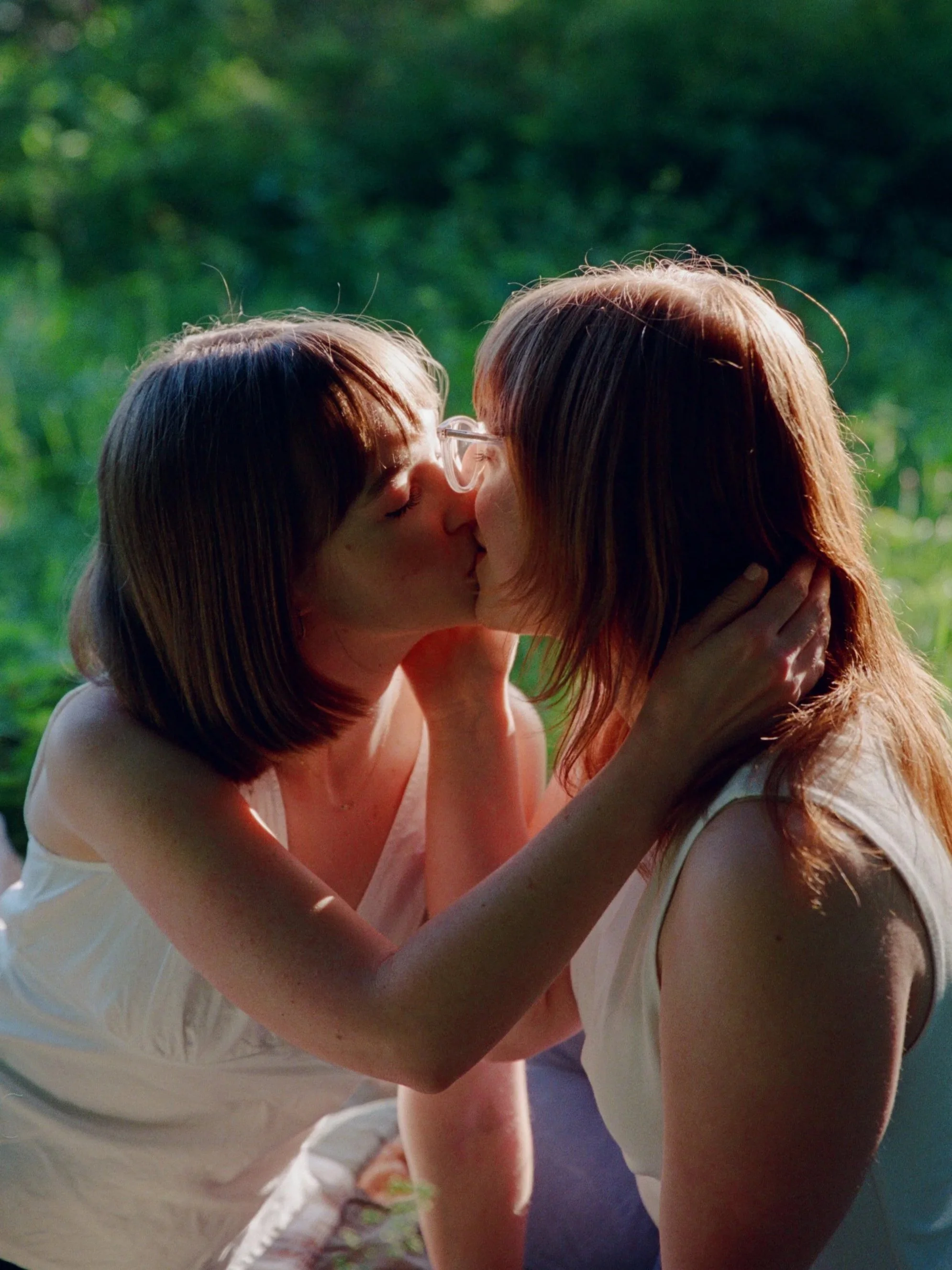 Two women with shoulder-length hair and light skin sharing a kiss outdoors with a natural green background. Shot by queer photographer Holly Stevens.