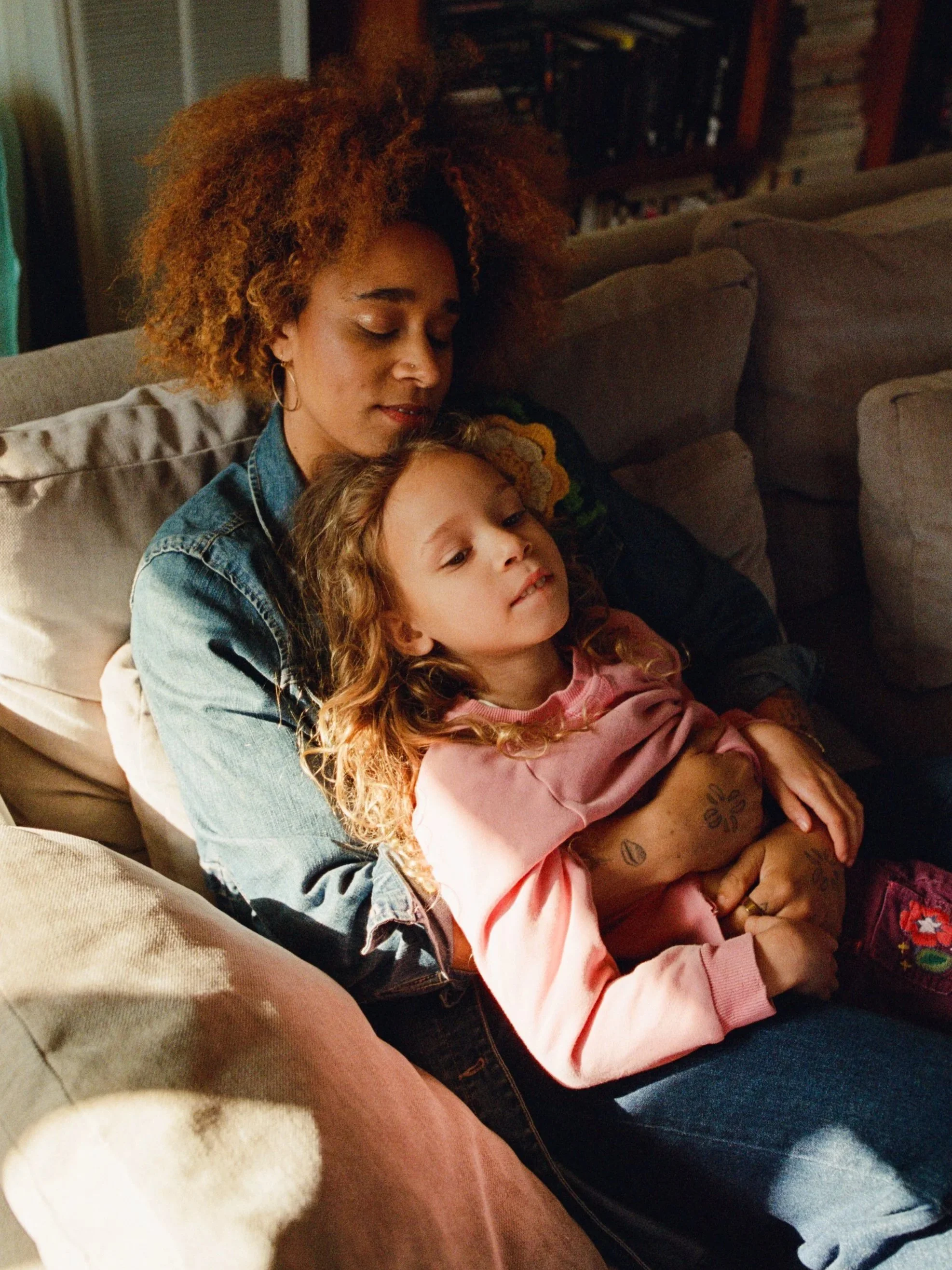 A woman with curly red hair and a young girl with curly hair sitting together on a beige couch. The woman has her eyes closed and the girl is lying against her, both appearing relaxed and content. Shot by Seattle photographer Holly Stevens.