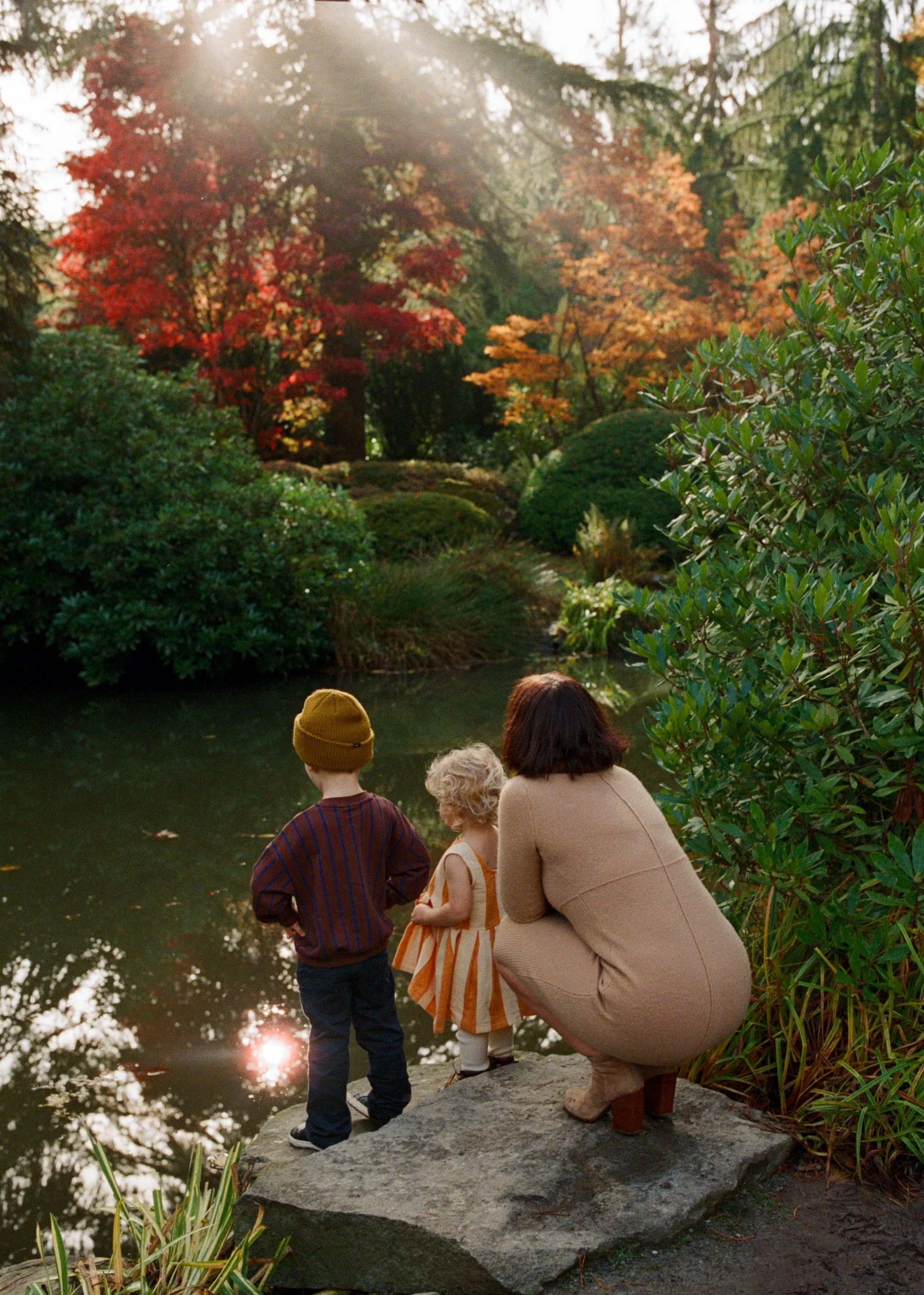 A woman with two young children, a boy and a girl, sitting on a rock by a pond in a park with autumn-colored trees and sunlight filtering through the branches.