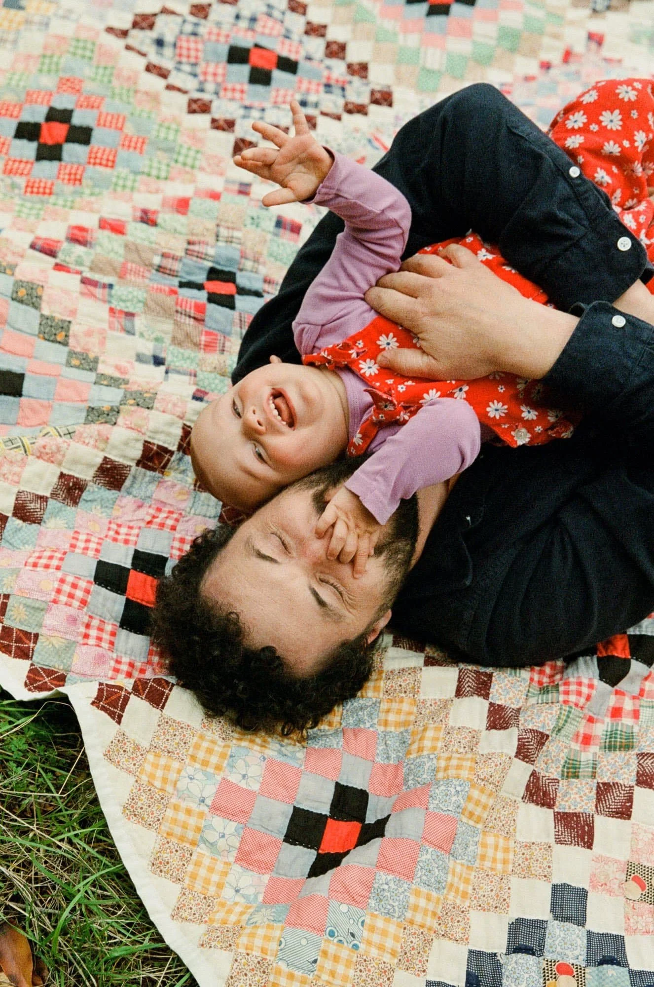 A man and a young girl lying on a colorful quilt outdoors. The girl is laughing and playfully tugging the man's nose, while the man has his eyes closed and appears to be enjoying the moment.