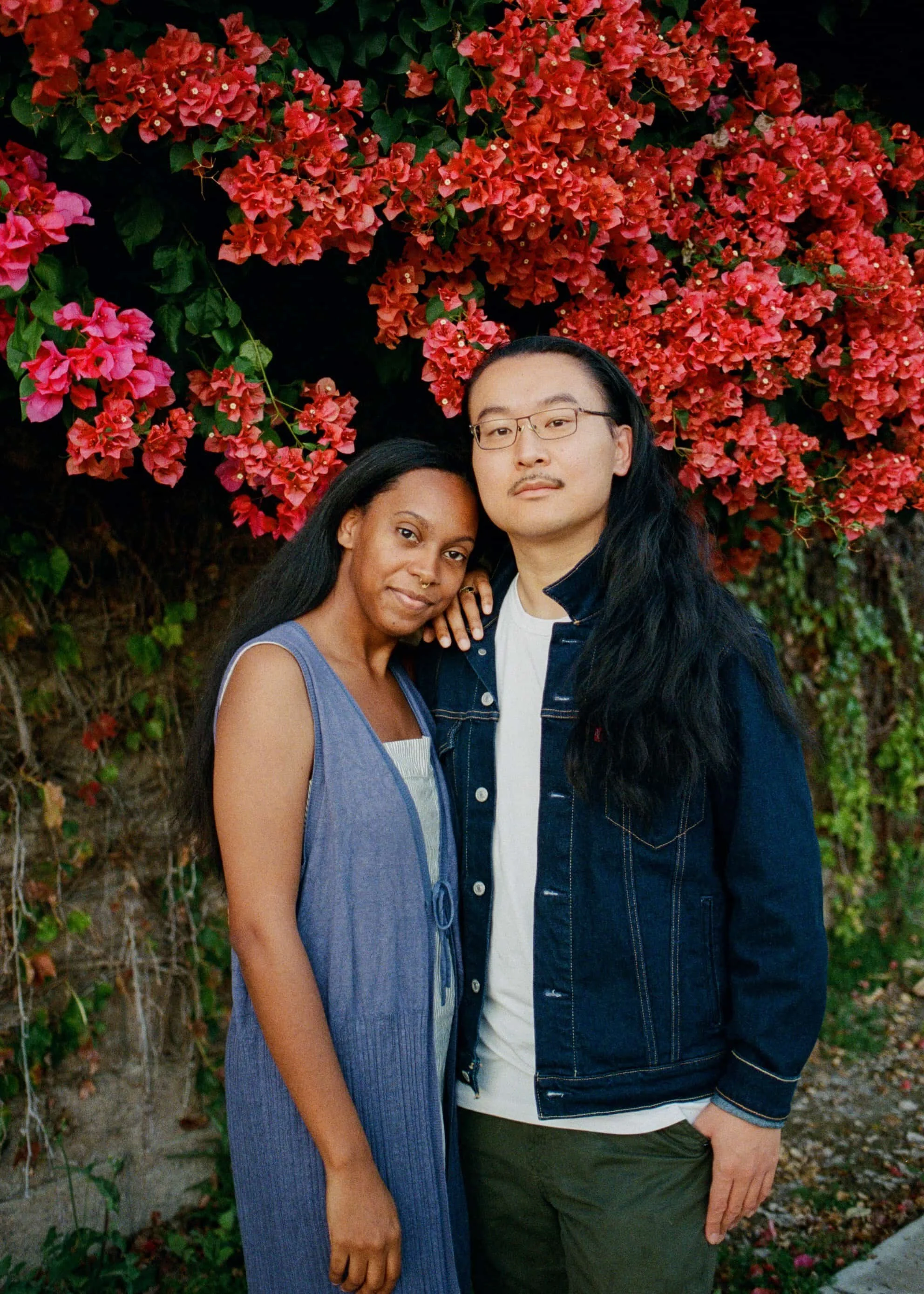 A man and woman standing close together in front of a vibrant pink and red bougainvillea flowers, with the woman resting her arm on the man's shoulder.