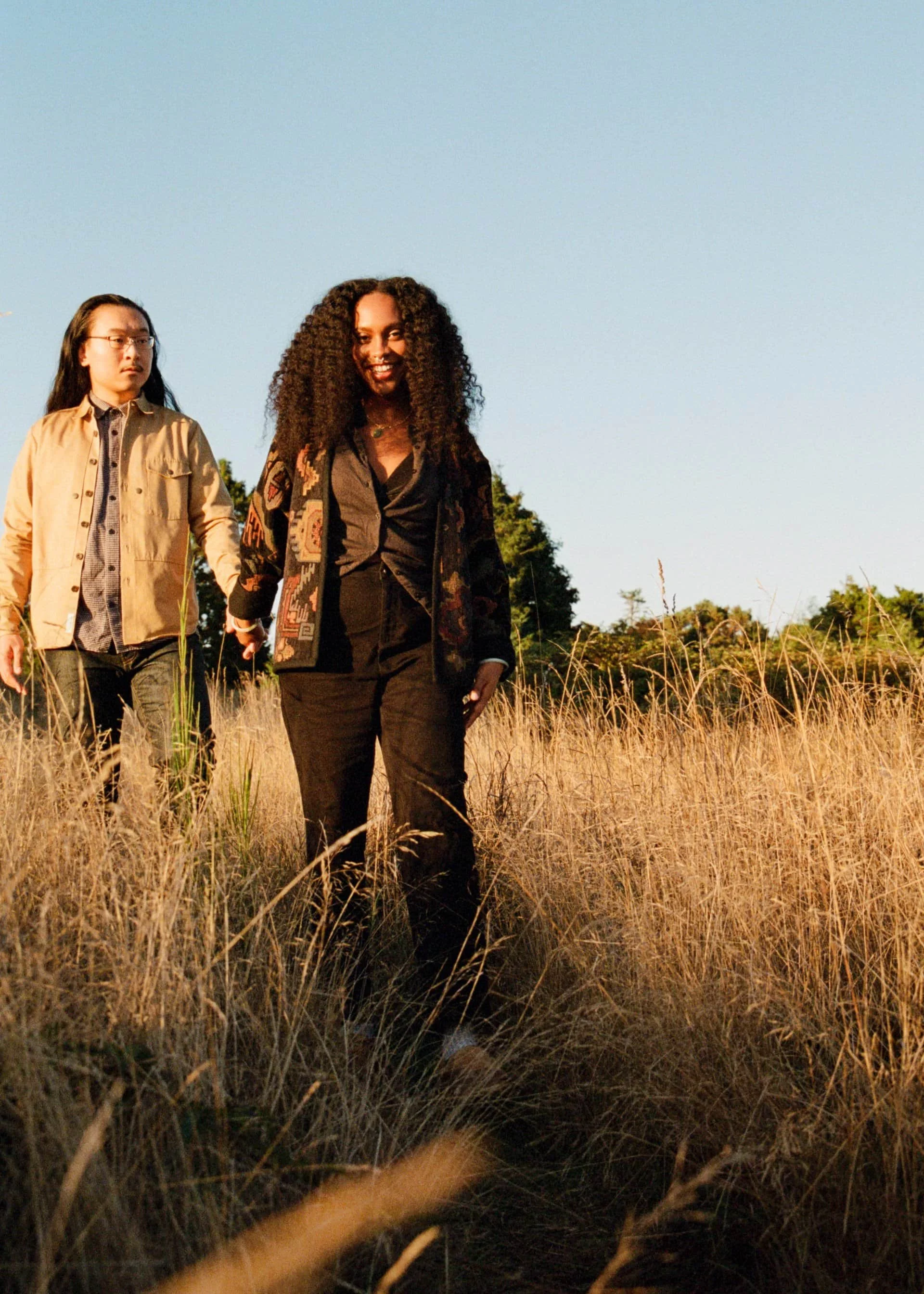 A man and a woman walking through a field of tall, dry grass during sunset, with clear blue sky and trees in the background.