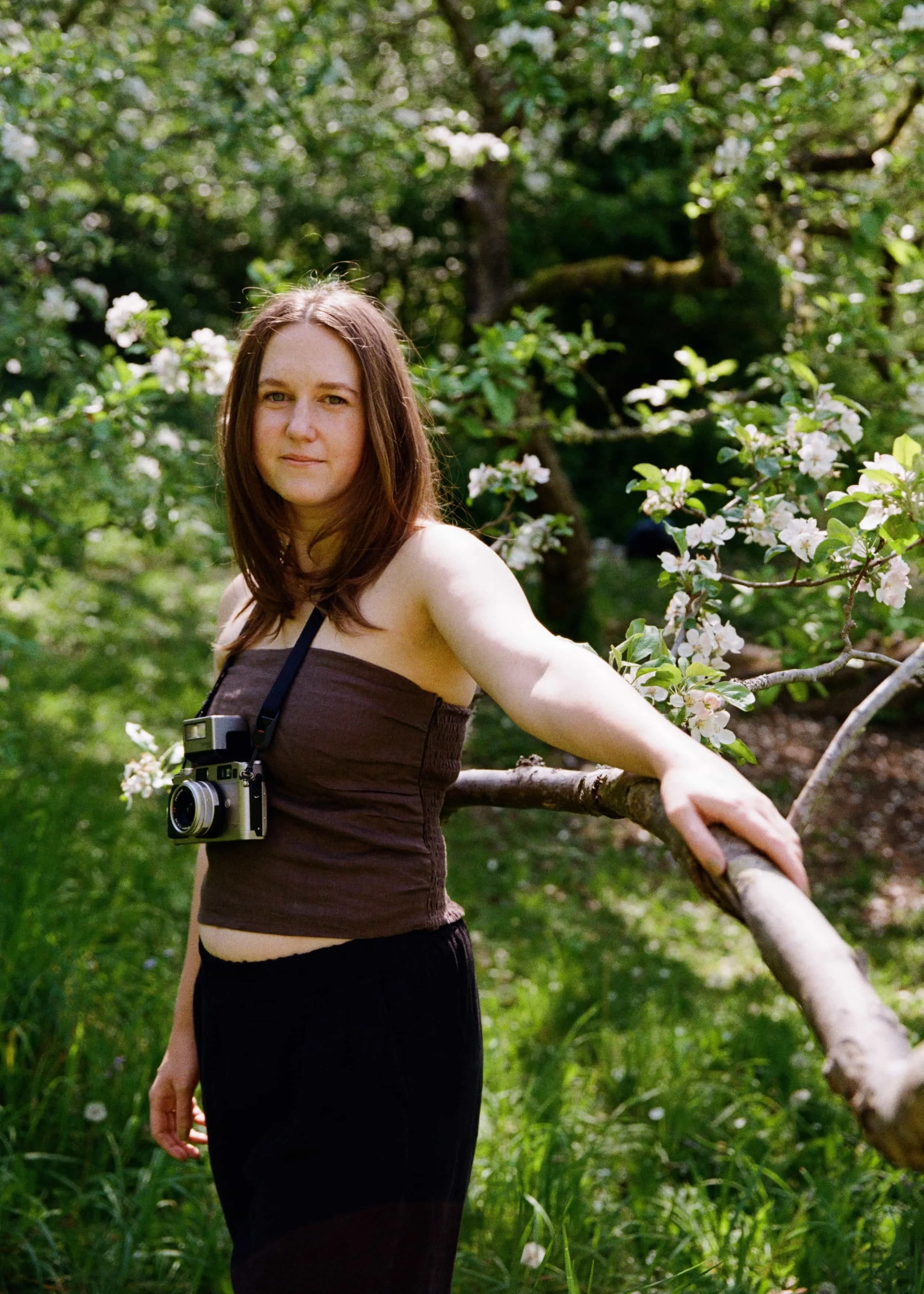 A woman leaning against a tree looking into the camera, with a slight smile on her lips and a camera hanging around her neck.