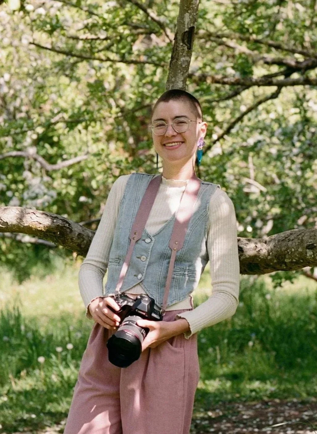 A nonbinary photographer in Seattle looking into the camera smiling, with a camera around their neck and their hands holding the camera.
